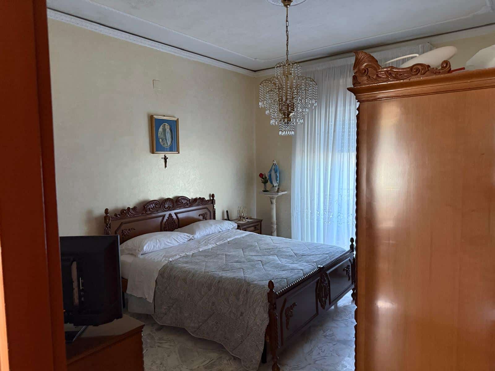 Letto matrimoniale in camera arredata in stile classico con mobili in legno intagliato, lampadario in cristallo e decorazioni religiose, ambiente accogliente e tradizionale.