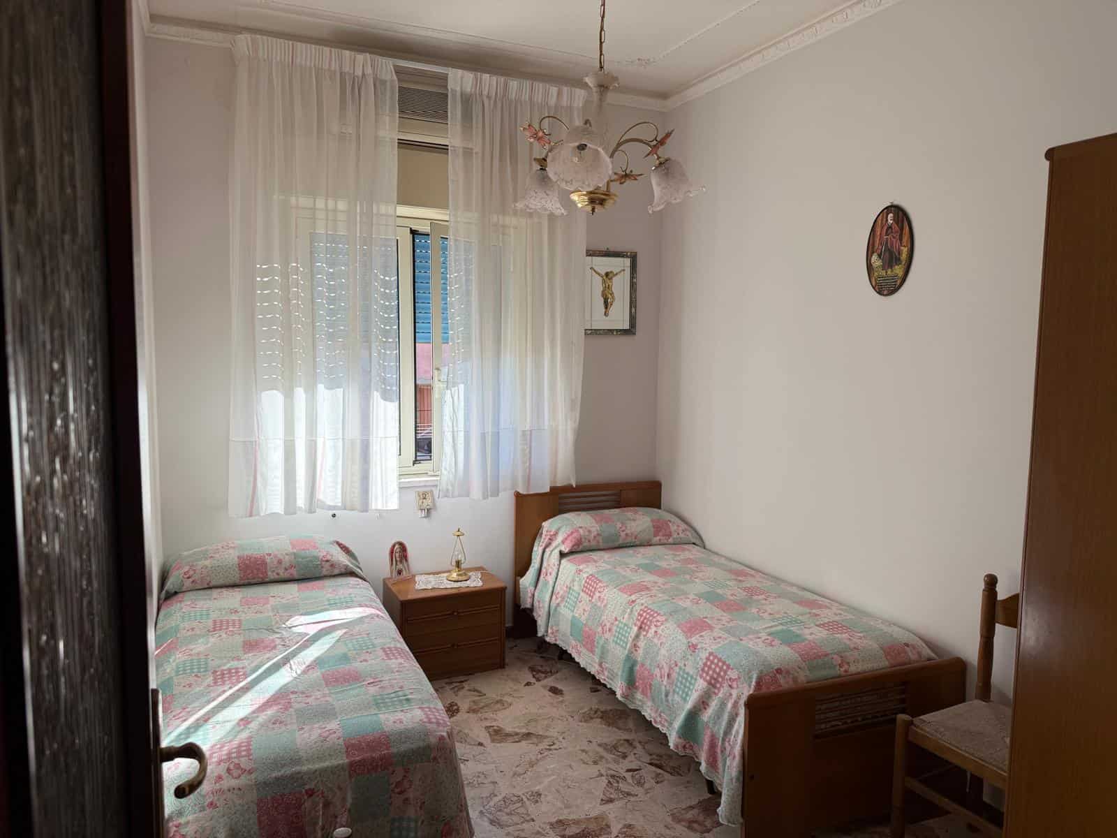 Letto singolo e lettino singolo in camera da letto con tende trasparenti e pareti bianche, decorazioni religiose e lampadario vintage, arredo in stile classico, ambiente accogliente e luminoso.