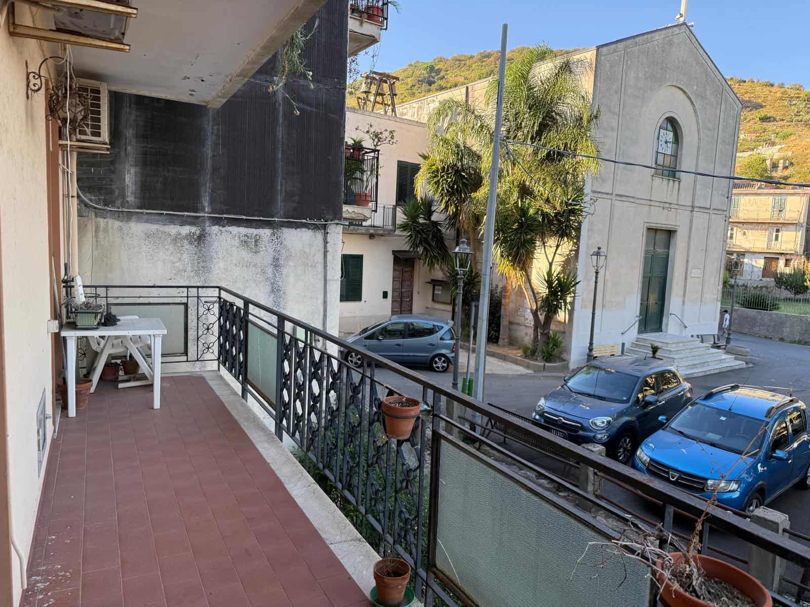 Balcone con vista su strada e chiesa, con piante in vaso e tavolo bianco, in un contesto di abitazioni e auto parcheggiate, atmosfera tranquilla e caratteristica di zona residenziale italiana.