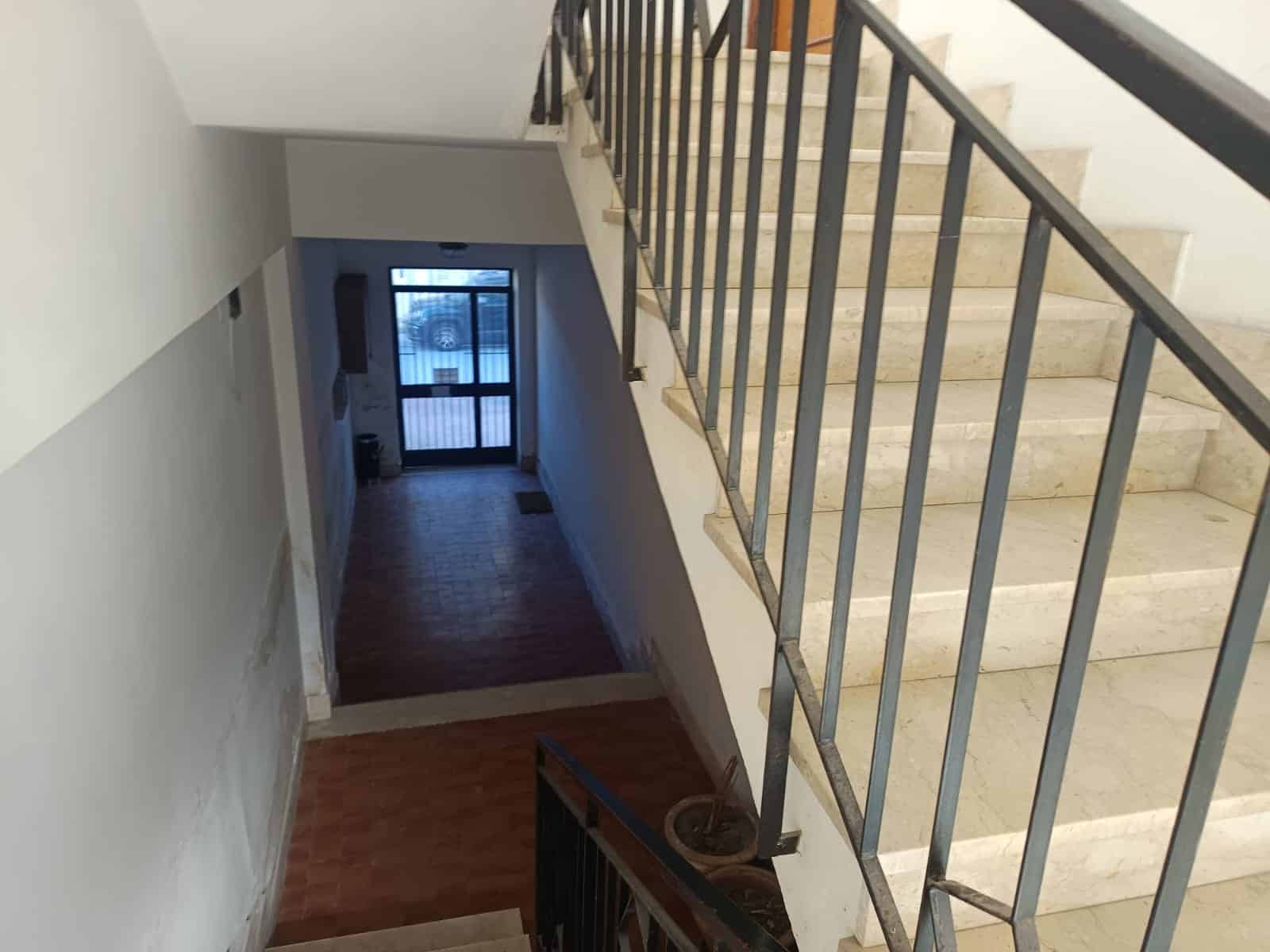 Scale condominiali in resina beige, corridoio luminoso con portone di ingresso in vetro e serranda motorizzata, ideale per immobili residenziali e commerciali.