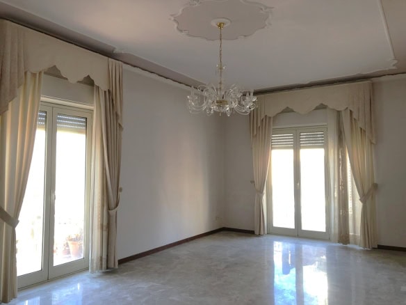 Finestre doppie con tende beige in stile classico, soggiorno luminoso con pavimento in marmo, soffitto decorato e lampadario elegante per un ambiente raffinato e spazioso.