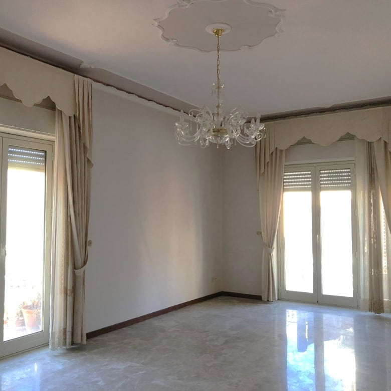 Finestre doppie con tende beige in stile classico, soggiorno luminoso con pavimento in marmo, soffitto decorato e lampadario elegante per un ambiente raffinato e spazioso.
