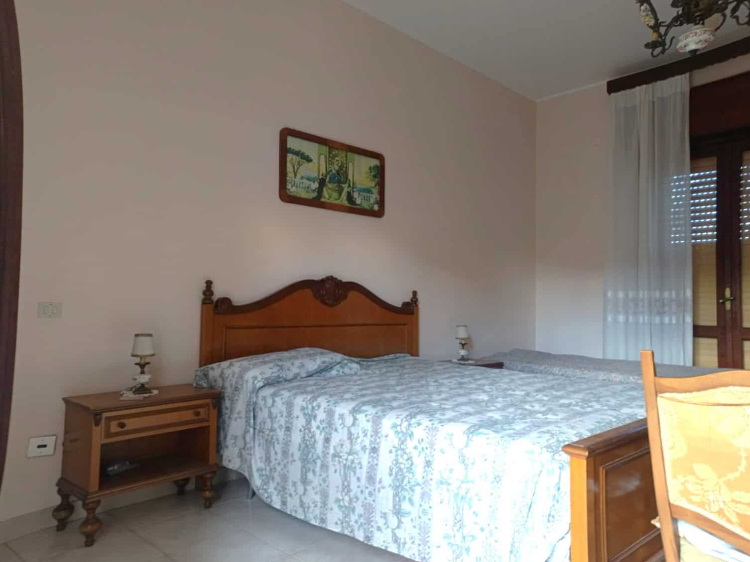 Letto matrimoniale in camera da letto con arredi classici, tapparelle e quadri decorativi, ambiente luminoso e accogliente, ideale per vivere in comfort e stile.