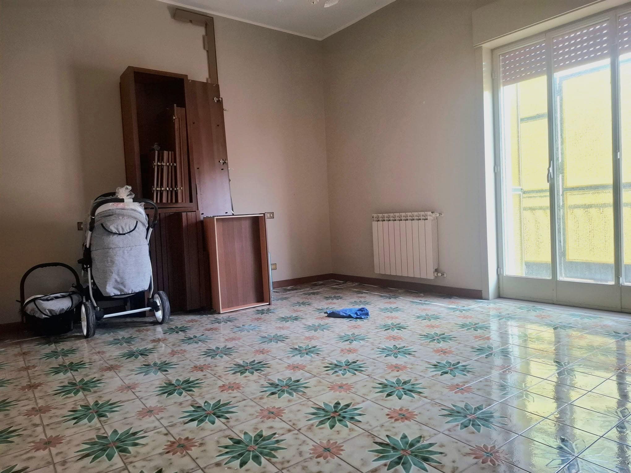 Aggiungi un'immagine di una stanza vuota con pavimento in piastrelle decorate, pareti beige, una finestra grande con persiane gialle e un radiatore.