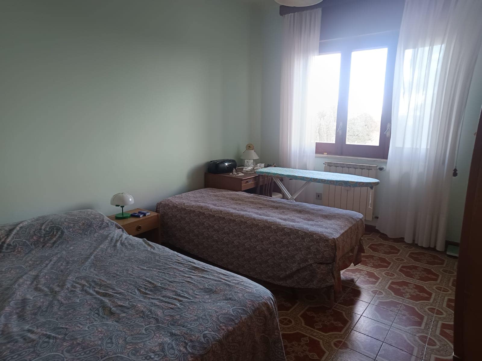 Letto matrimoniale e letto singolo in camera da letto con finestra luminosa, arredi classici e pavimento in ceramica decorata, ideale per affitto case o residenti.