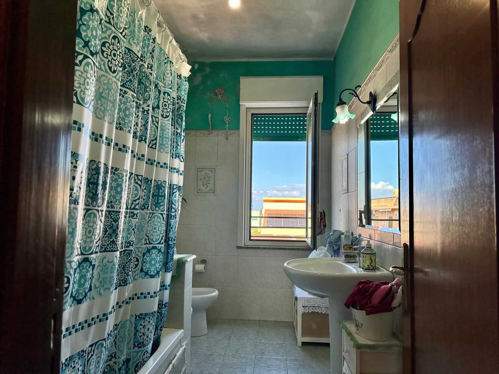 Vaschetta da bagno con tenda trasparente blu e bianca, finestra che lascia entrare molta luce naturale, vista aperta sulla città, arredo bagno tradizionale, dettagli di arredo e decorazioni.
