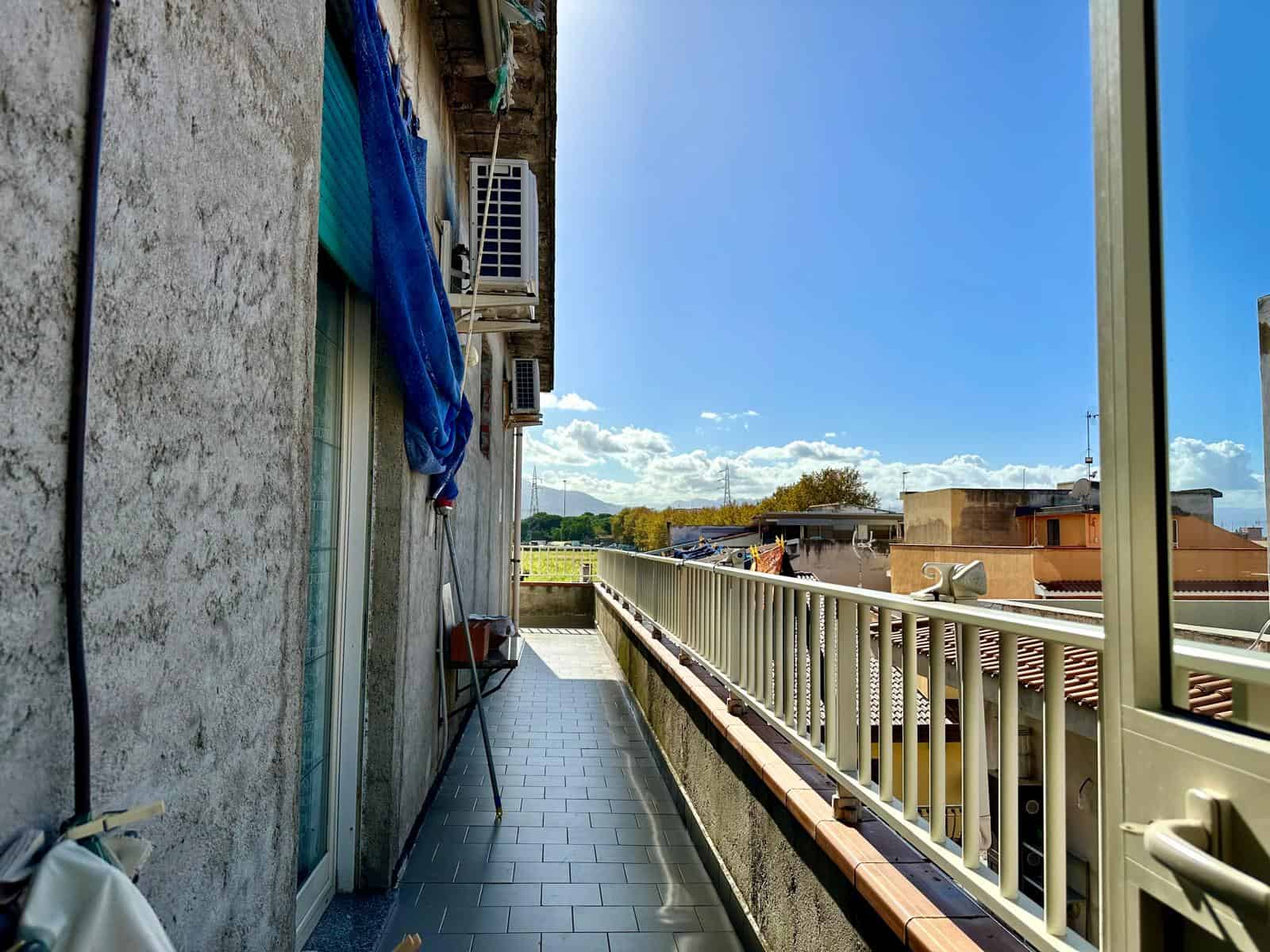 Balcone di un appartamento con cielo blu, parasole, tende, aria condizionata e viste su tetti e paesaggio naturale, ideale per relax e divertimento all'aperto.