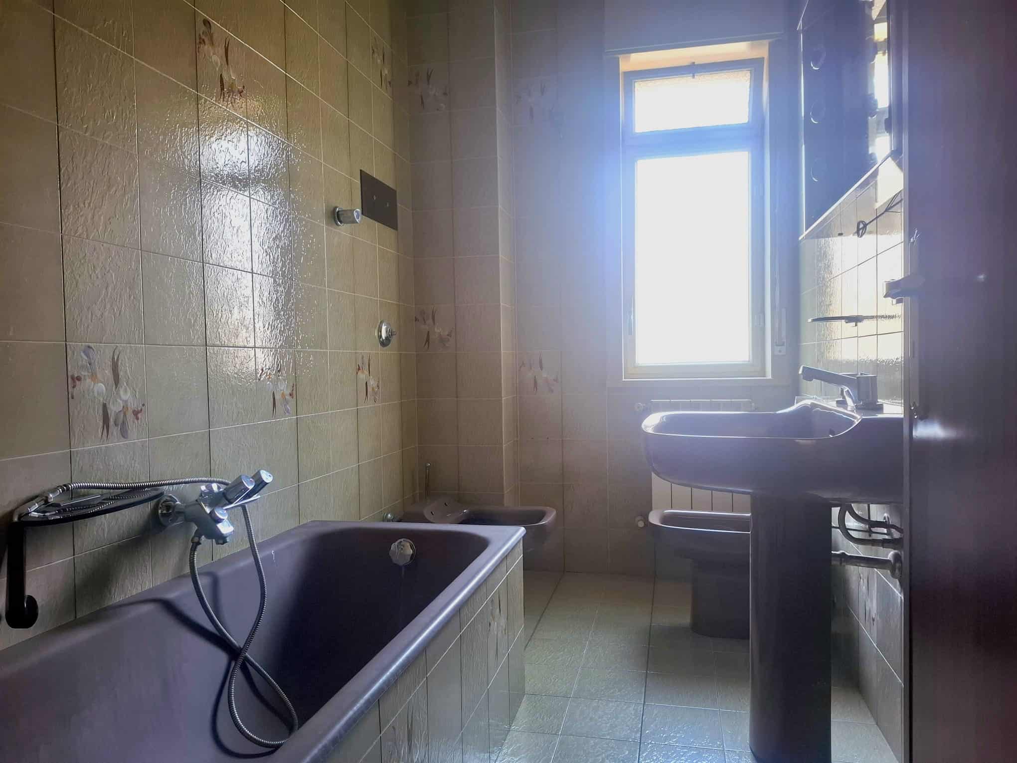 Lavabo e vasca da bagno in un bagno di vecchio stile con piastrelle beige, finestra luminosa e arredi essenziali, ideale per ristrutturazioni o rinnovamenti di case.