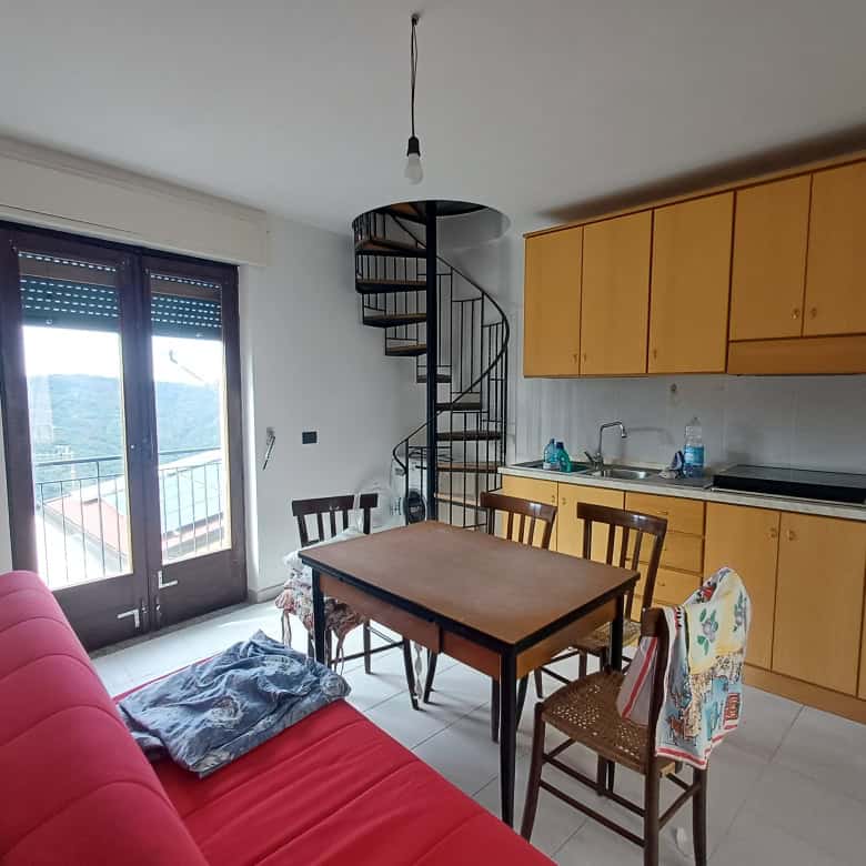Spazio soggiorno con cucina a vista, balcone con vista panoramica, arredi moderni, area soggiorno con divano rosso, tavolo in legno, scale a spirale in metallo, aria condizionata, ambiente luminoso e accogliente.