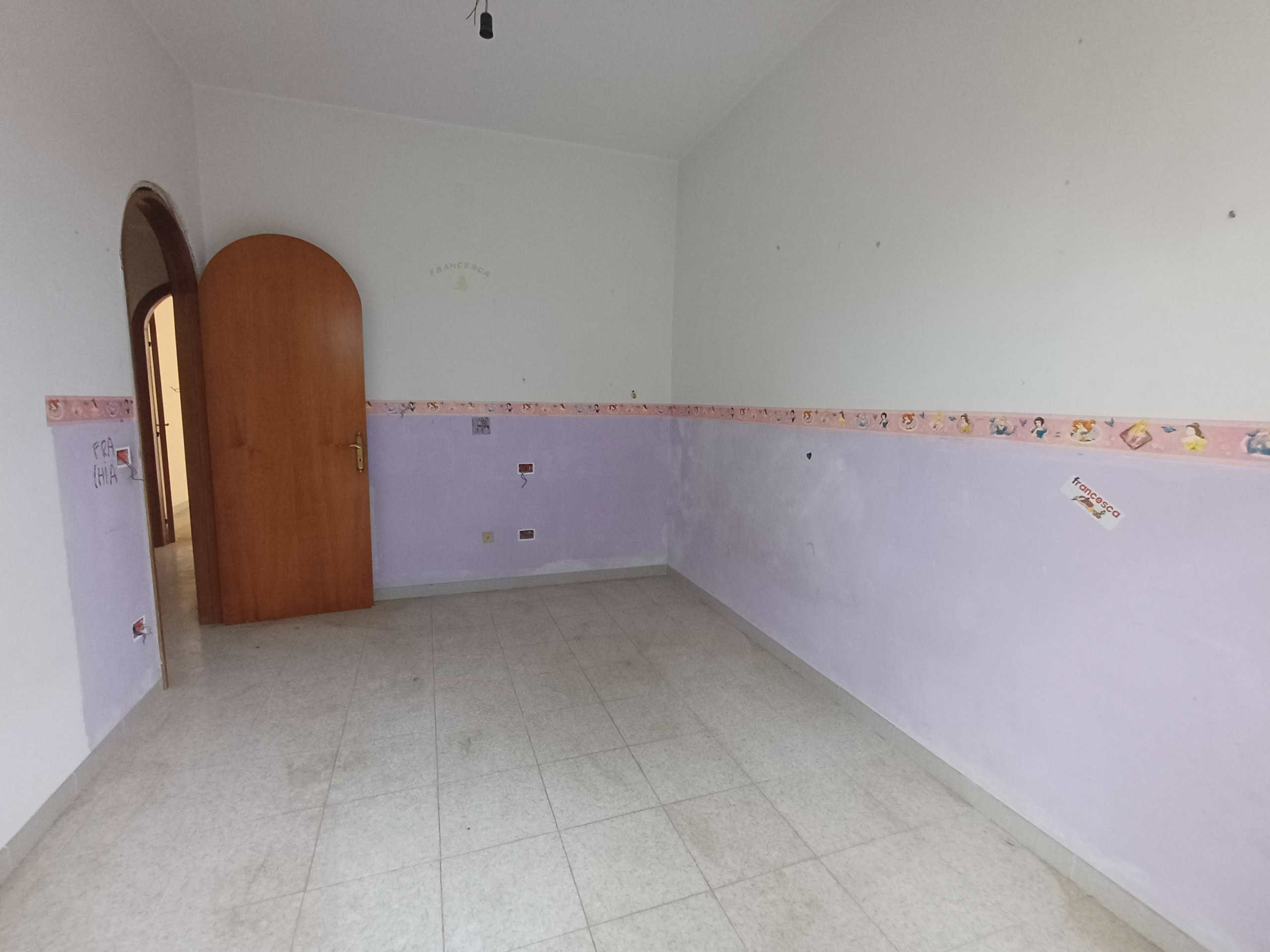 Prima casa da ristrutturare, stanza vuota con pareti lilla e soffitto bianco, pavimento in piastrelle beige, porta in legno, dettagli adesivi decorativi e fotografie. Ideale per investimento immobiliare.