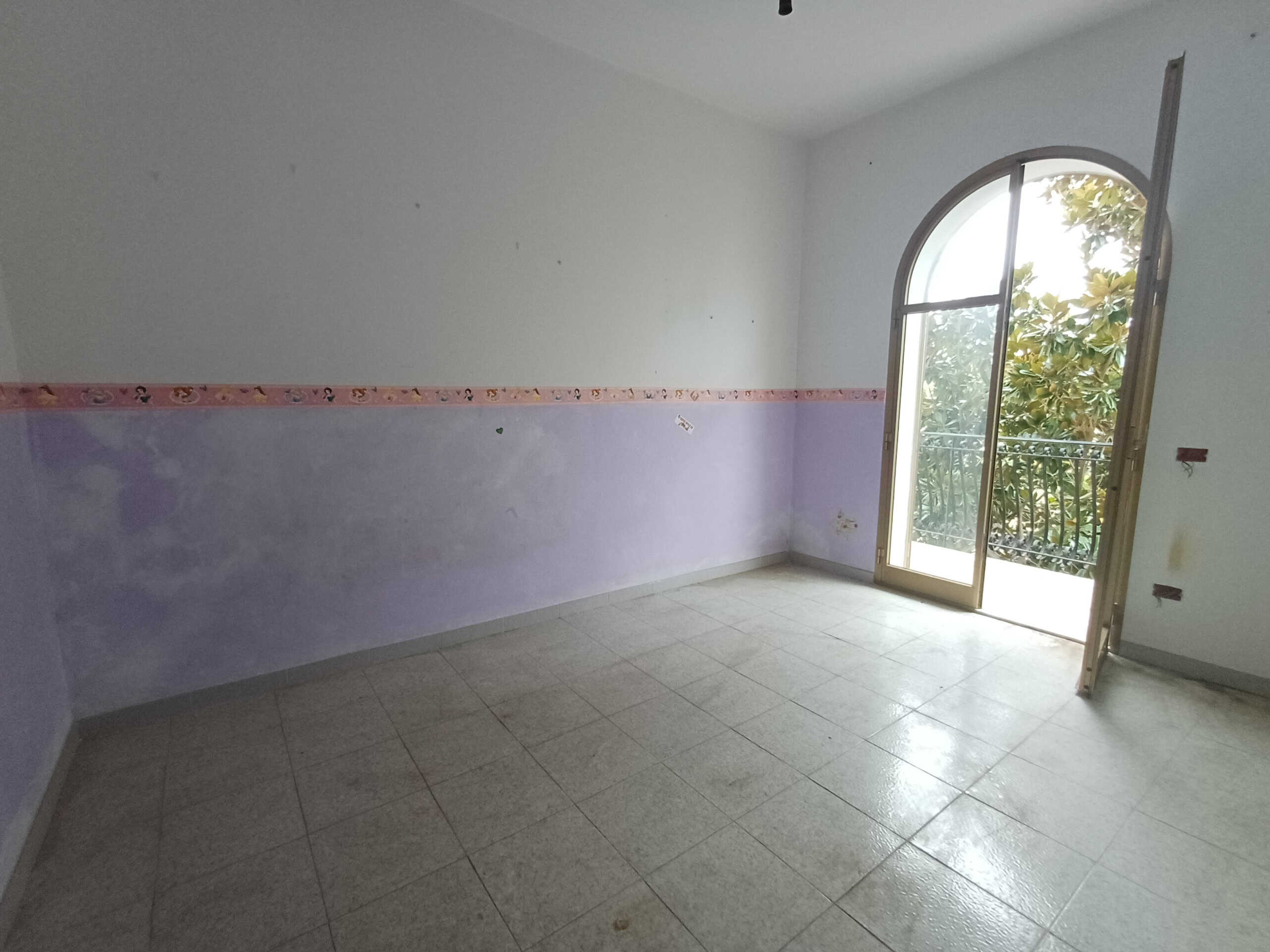 Spazio vuoto per camera da letto con finestra ad arco e balcone, pareti color lavanda, pavimento in piastrelle chiare, ideale per ristrutturazione e arredamento personalizzato.