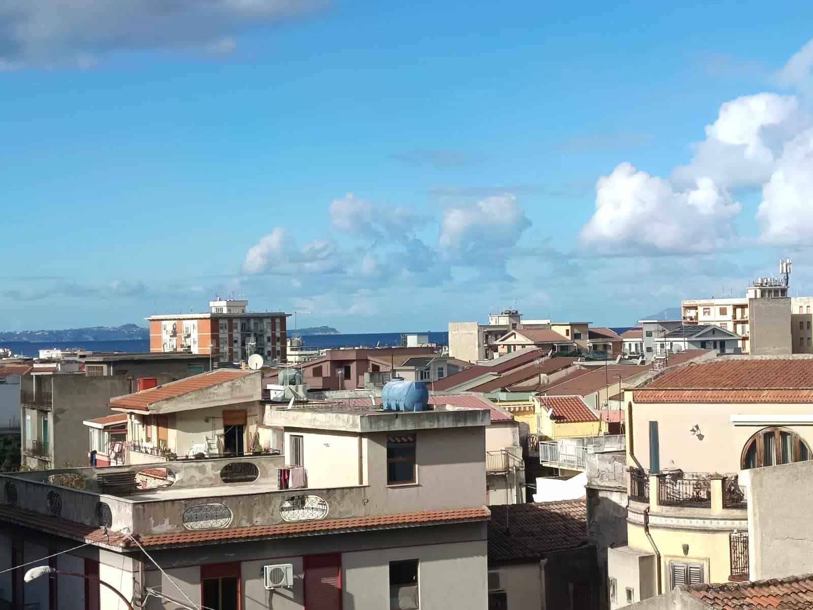 Vista panoramica sul tetto di case e palazzi con il mare in lontananza sotto un cielo azzurro con nuvole bianche, scenario ideale per immobili e investimenti immobiliari.