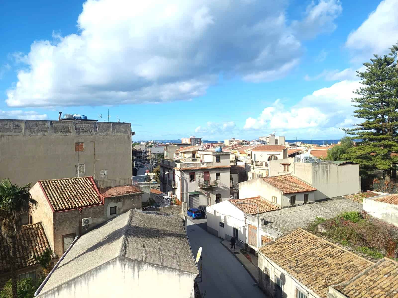 Panorama urbano con tetti in tegole, cielo sereno e nuvole bianche, vista sul mare, edifici residenziali, strada tranquilla e natura, perfetta per annunci immobiliari e promozione di case vicino al mare.