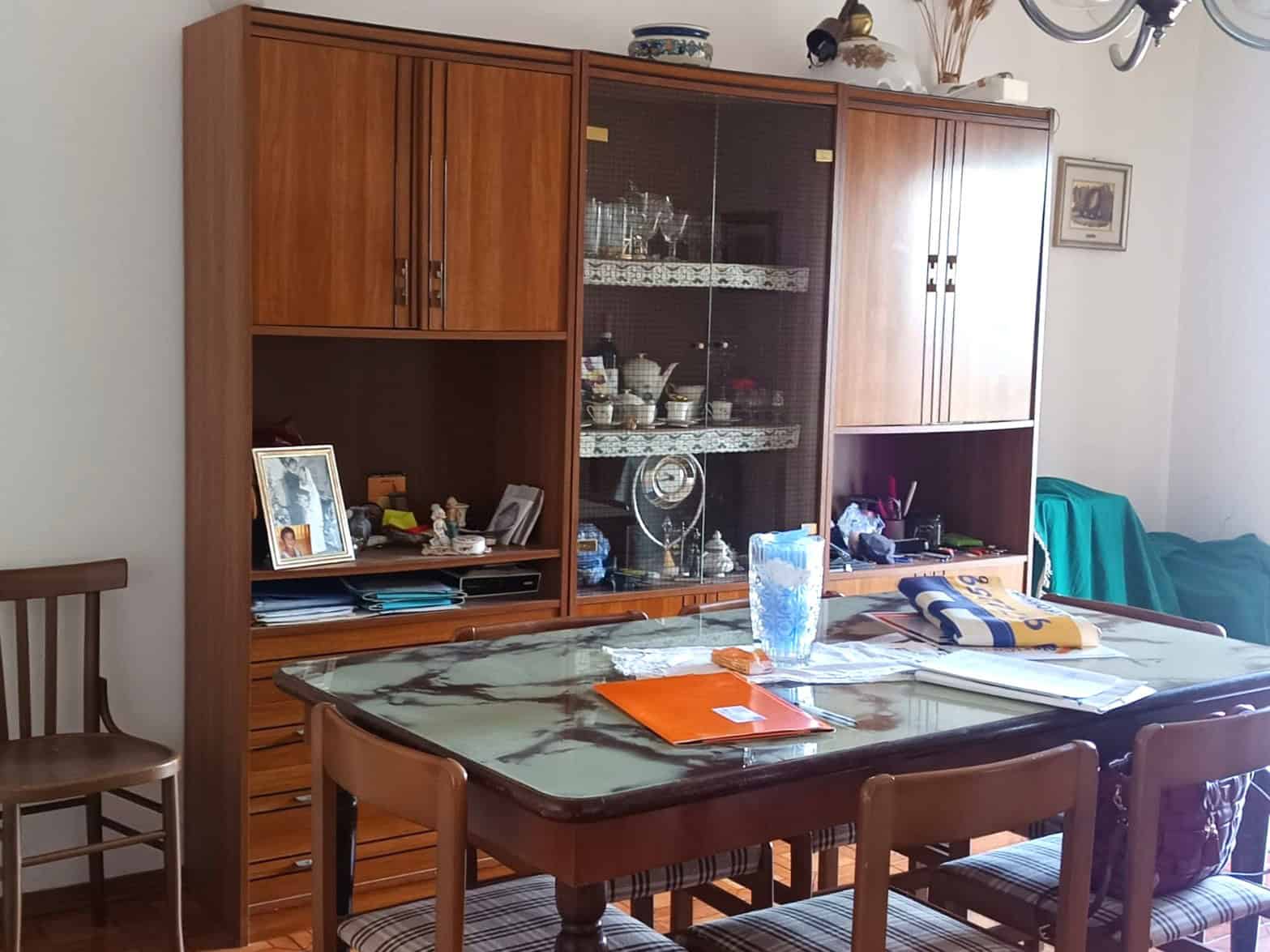 Mobili soggiorno in stile classico, credenza in legno con vetrinetta con articoli in porcellana e cristallo, tavolo con sedie, decorazioni e oggetti domestici.