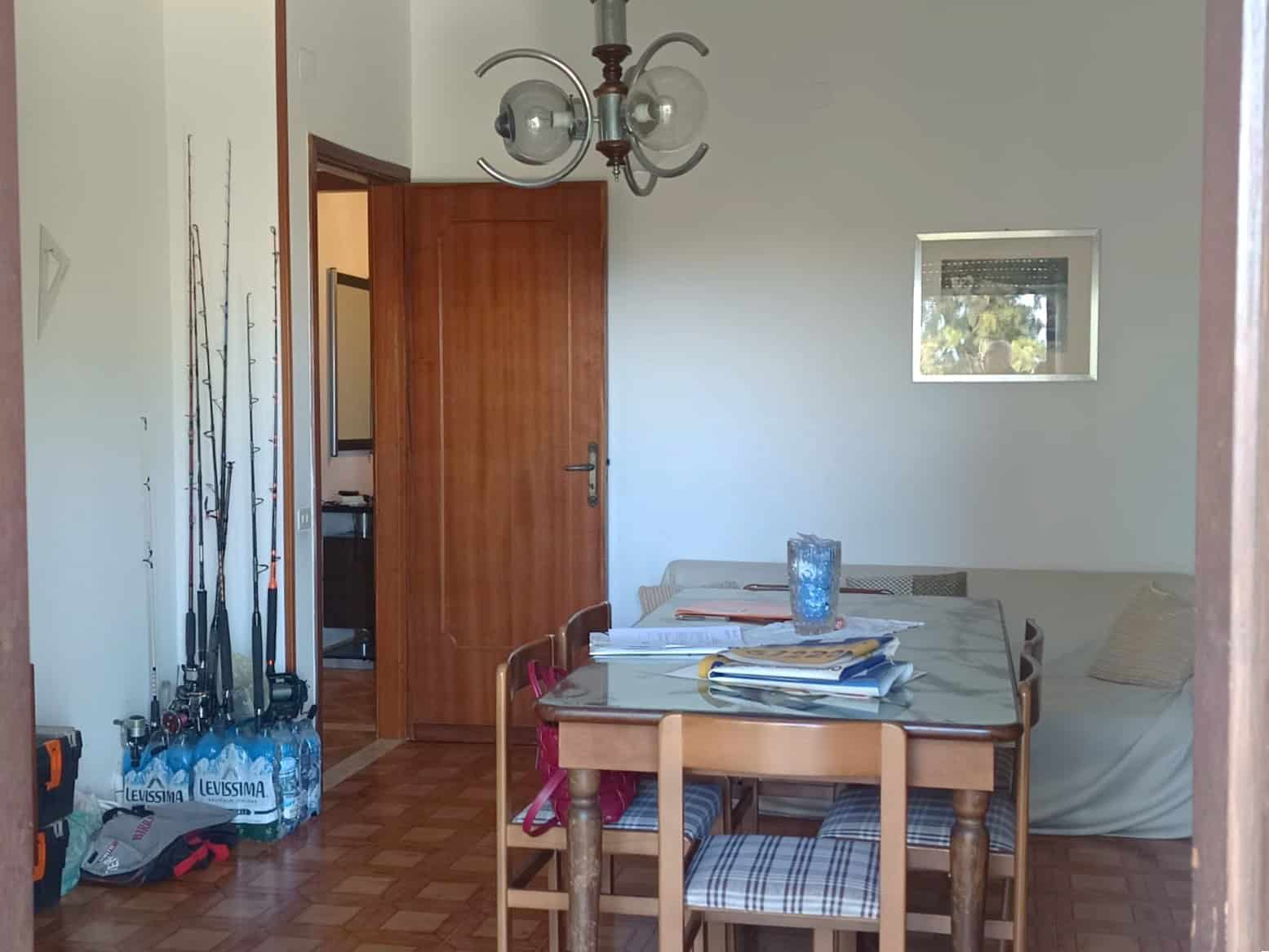 Mobili soggiorno con tavolo e sedie in legno, parete bianca con piccola finestra e lampadario vintage; ambiente domestico accogliente e funzionale.