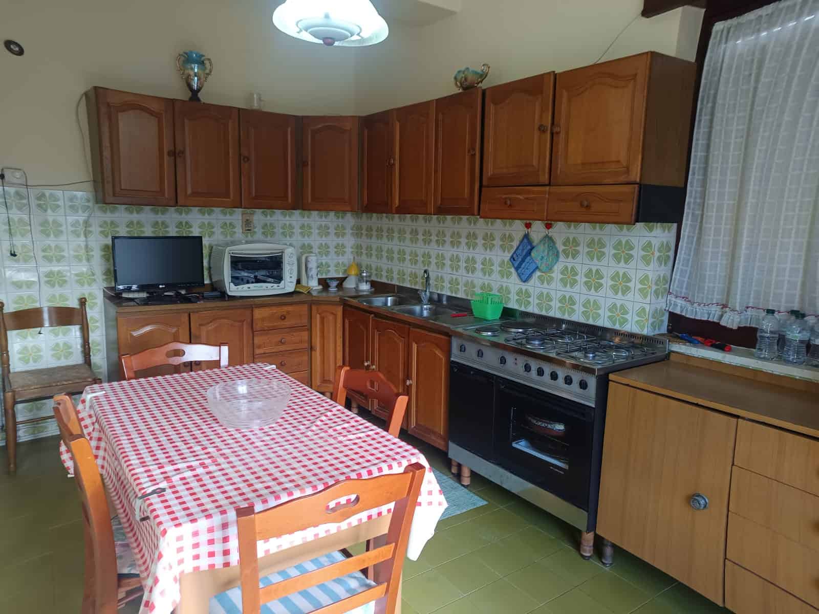 Lavagna cucina con mobili in legno, piano cottura a gas, tavolo con tovaglia a scacchi rossi e bianchi, macchina per il pane, tv e utensili vari, ambiente accogliente e funzionale.