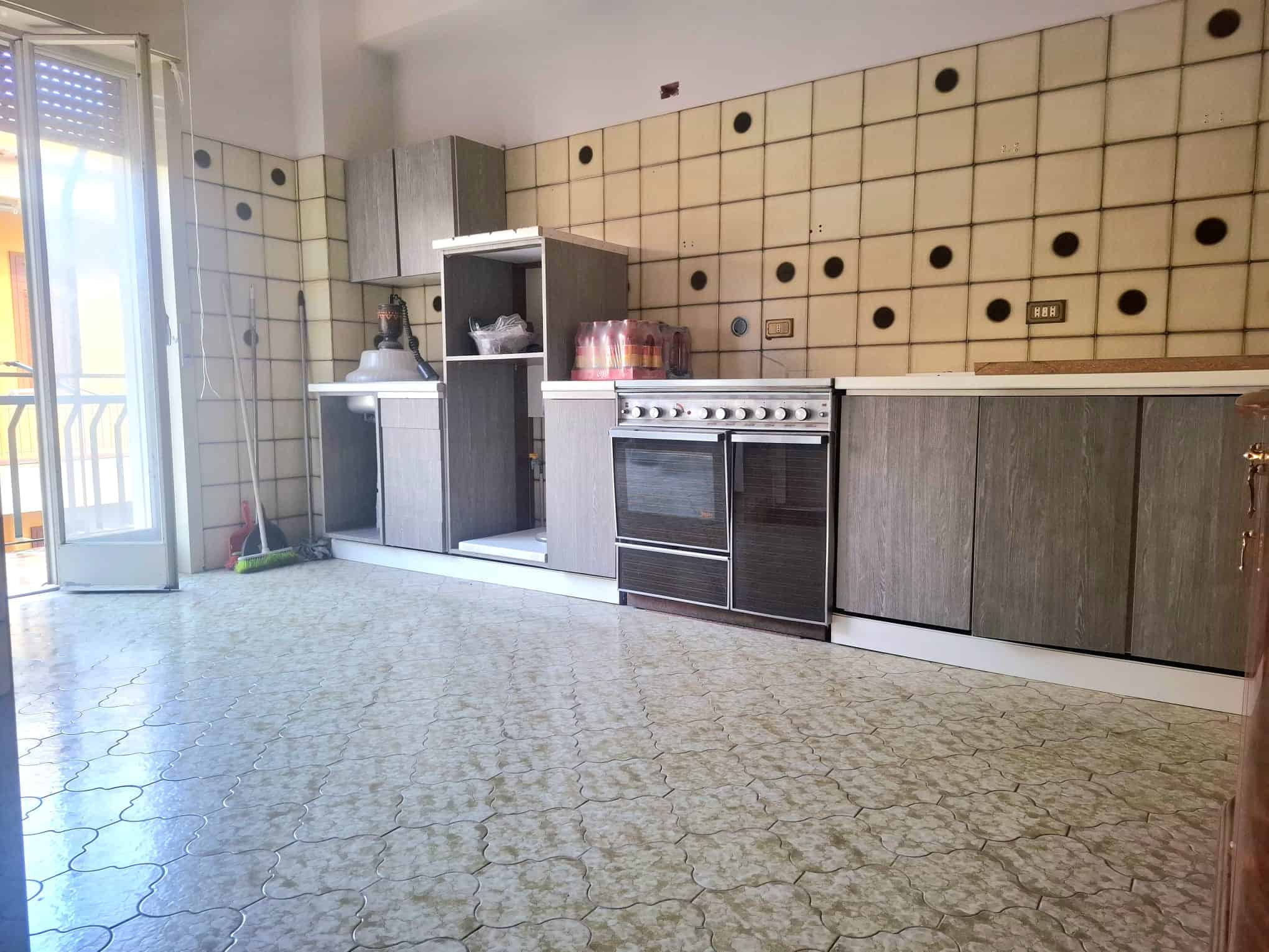 Pannello di cucina moderna con mobili in legno grigio e parete decorativa con piastrelle beige e nero, spazio luminoso con accesso al balcone, ideale per ambienti abitativi contemporanei.