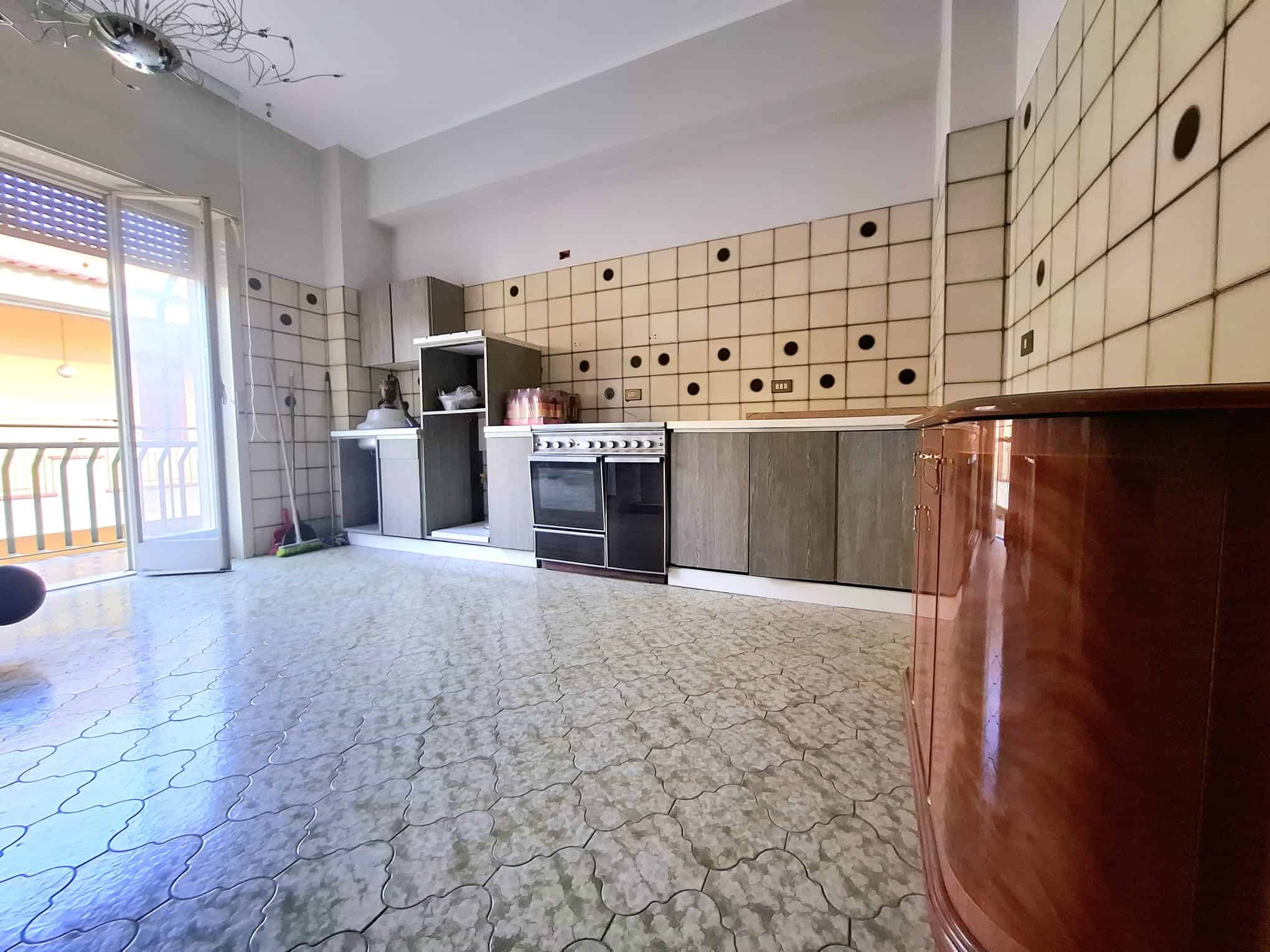 Ampio soggiorno con cucina moderna, pareti rivestite in piastrelle beige con dettagli neri, pavimento in marmo chiaro, balcone con vista luminosa, ideale per vivere e intrattenere.