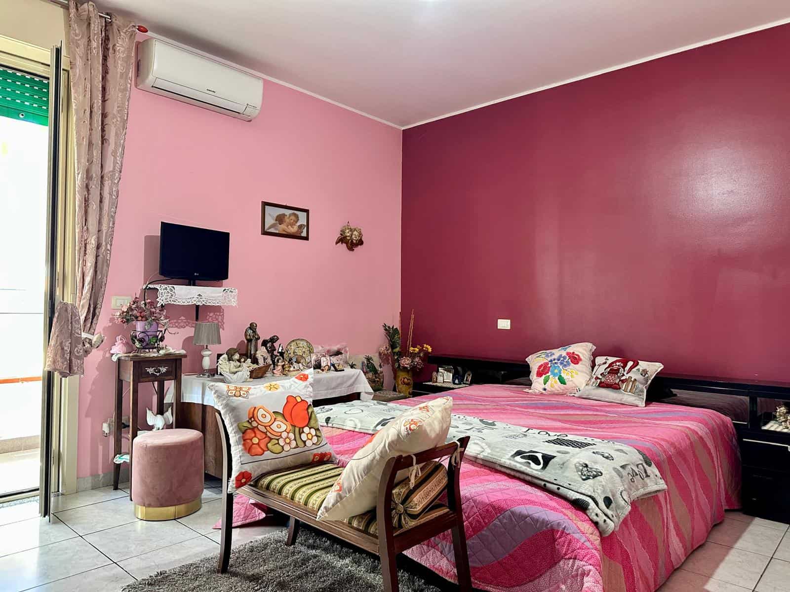 Letto matrimoniale con coperte rosa e cuscini decorativi in camera da letto colorata, arredi tradizionali e decorazioni religiose, stanza accogliente con pareti rosa pastello e mobili funzionali.