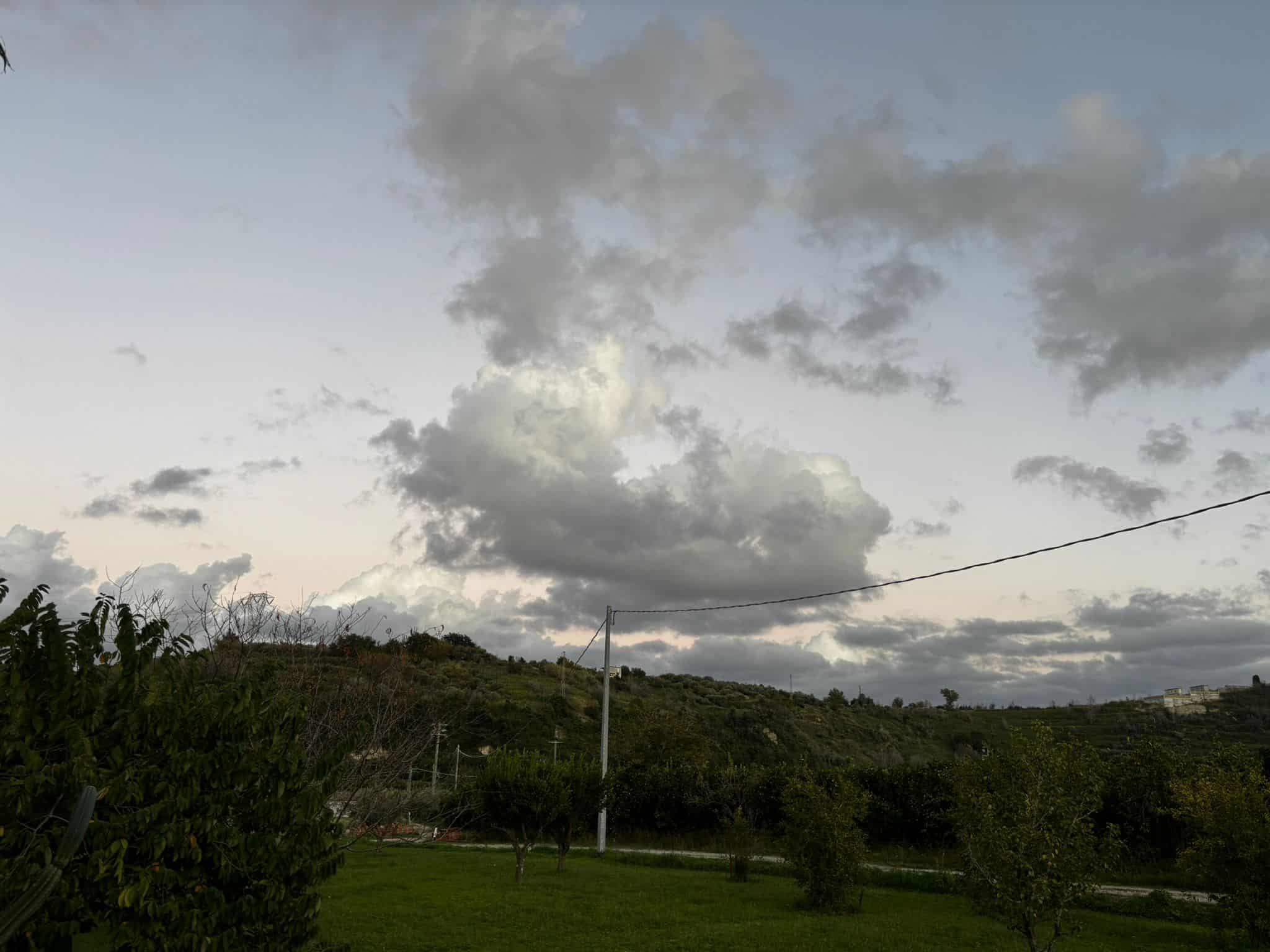 Nevicata invernale su una casa di campagna immersa nella natura, con alberi e colline verdi, cielo nuvoloso e atmosfera tranquilla e serena.