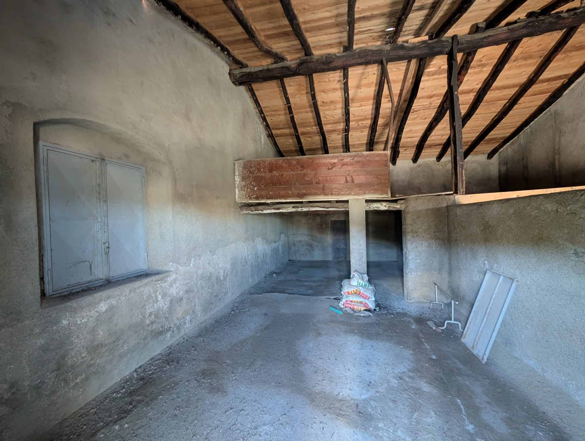 Vecchia soffitta in ristrutturazione, pareti in calcestruzzo, tetto in legno con travi a vista, spazio vuoto per futura conversione residenziale o commerciale, materiali da costruzione a terra.