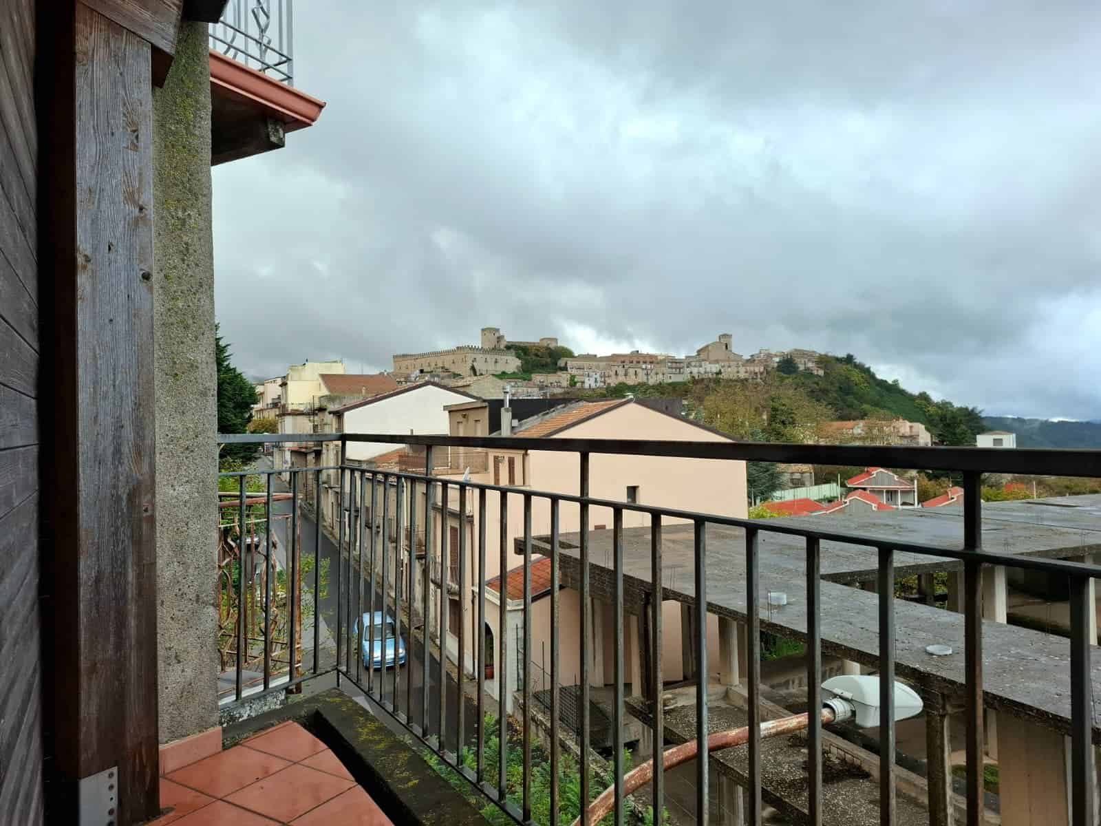 Vista panoramica su un antico castello situato in cima a una collina, vista dalla terrazza di un appartamento con arredi in legno e ferro battuto, con tetto in tegole rosse e strade pittoresche sottostanti.
