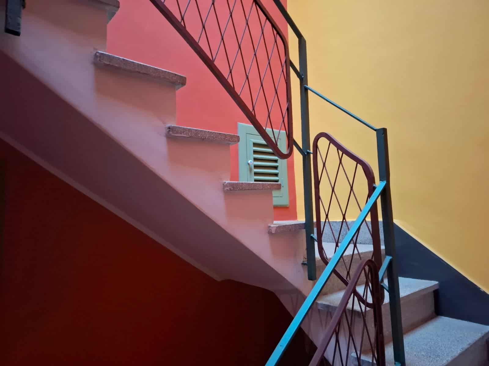 Scale di colore vivace con ringhiera decorativa in ferro battuto, uscita finestra con grate a scacchiera, pareti esterne dai colori caldi, ideale per appartamenti e case moderne o in stile mediterraneo.