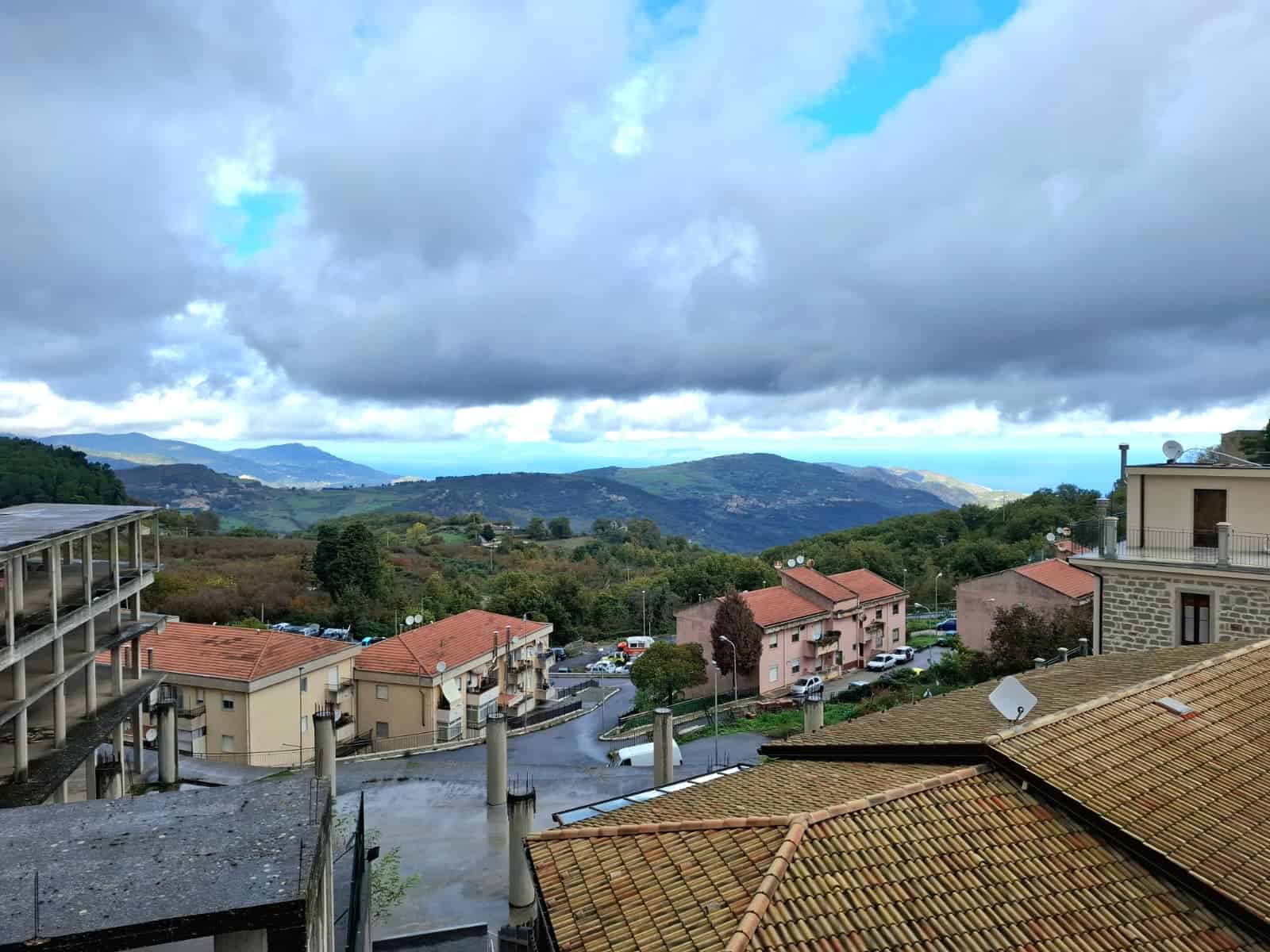 Valle panoramico con case in collina, cielo nuvoloso, vista mare in lontananza, case con tetti in scandole e terrazze, scenari rurali e paesaggio naturale.