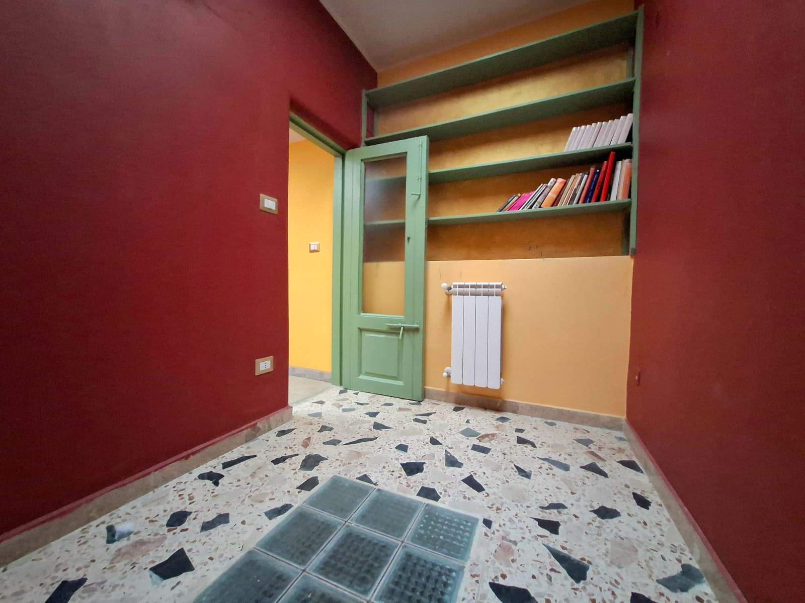 Ripostiglio colorato in stile vintage con scaffali verdi, pareti arancioni e rosse, pavimento in marmo con piastrelle colorate, ideale per la casa o l'ufficio.