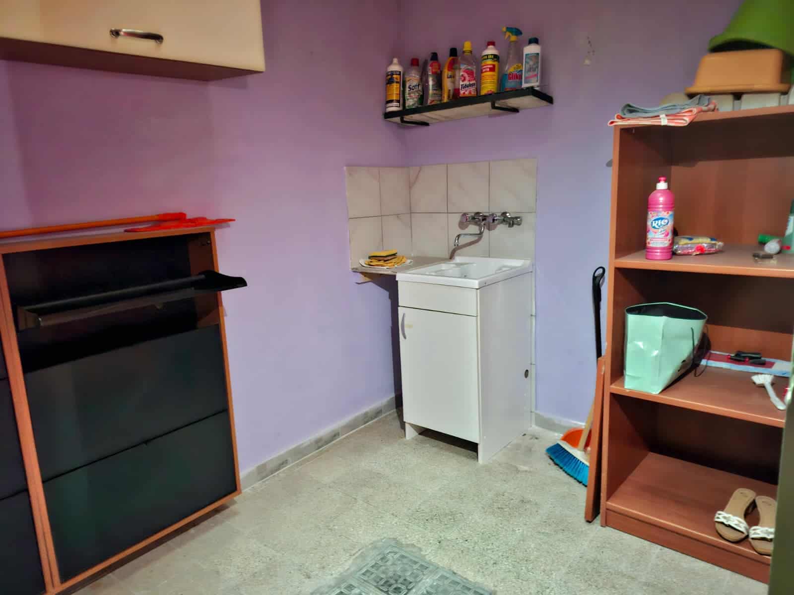 Lavanderia con lavabo, scaffali per detergenti e spazio per stendere, pareti lilla, pavimento in piastrelle beige, accessori per pulizia e scarpe, ambiente funzionale e pratico.