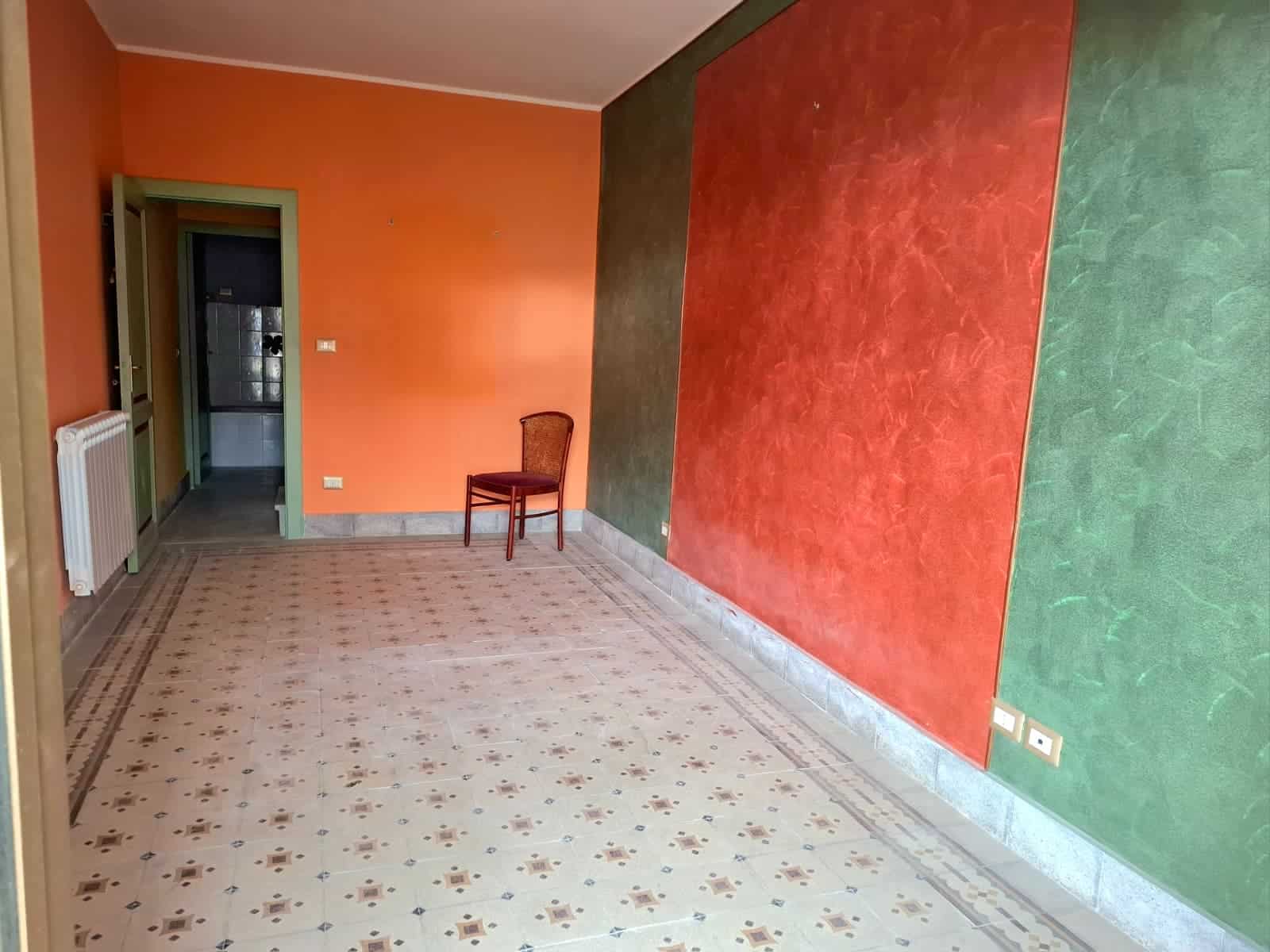 Soggiorno con pareti colorate in arancione, rosso e verde, pavimento in piastrelle decorative, e una sedia posizionata vicino alla parete verde, ambiente accogliente e vivace.