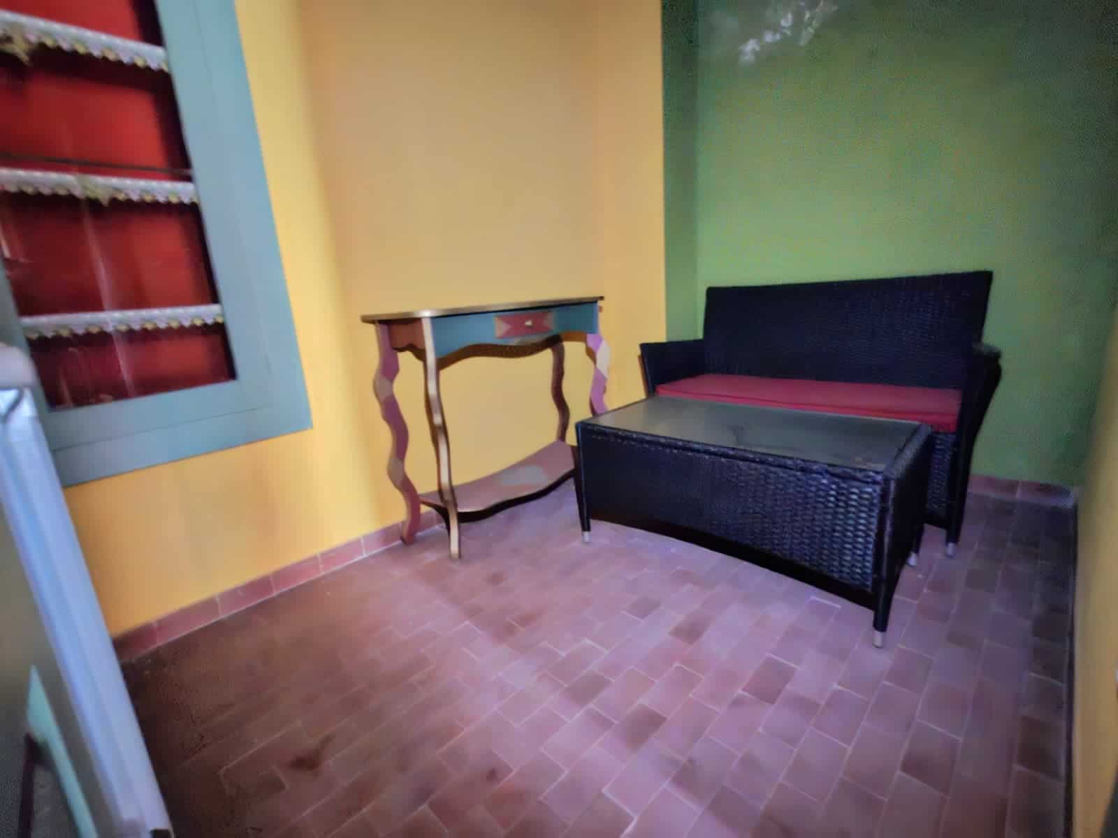 Spazio esterno di un'abitazione con pareti colorate, arredi in vimini nero e un'apertura con tenda con fantasie floreali, ideale per relax e momenti di convivialità.