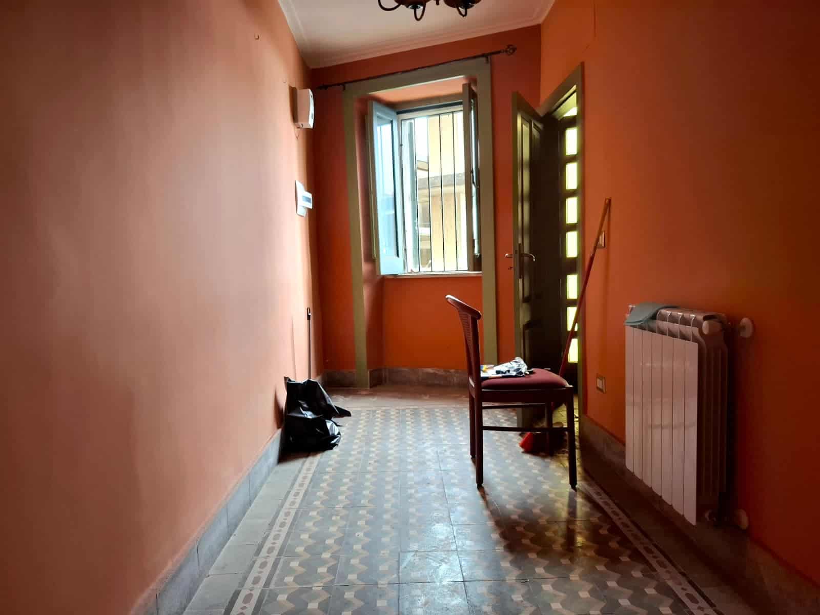 Serranda di una finestra aperta in una stanza con pareti arancioni e pavimento in gres decorato, con una sedia, un termosifone e oggetti vari, ambiente luminoso e accogliente.