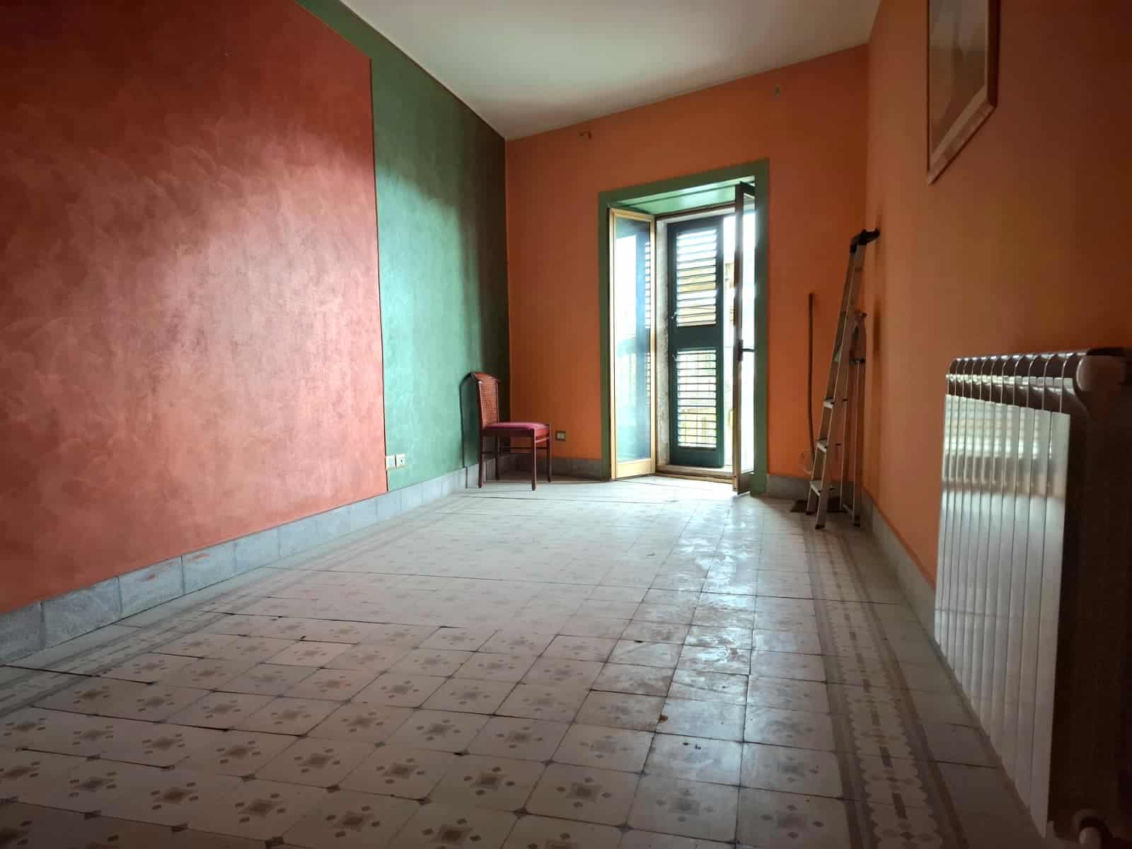 Spazio vuoto in appartamento con pareti colorate e porta finestra che dà sul balcone, arredato con sedia e scala, pavimento in ceramica, ideale per ristrutturazione o nuovo arredo.