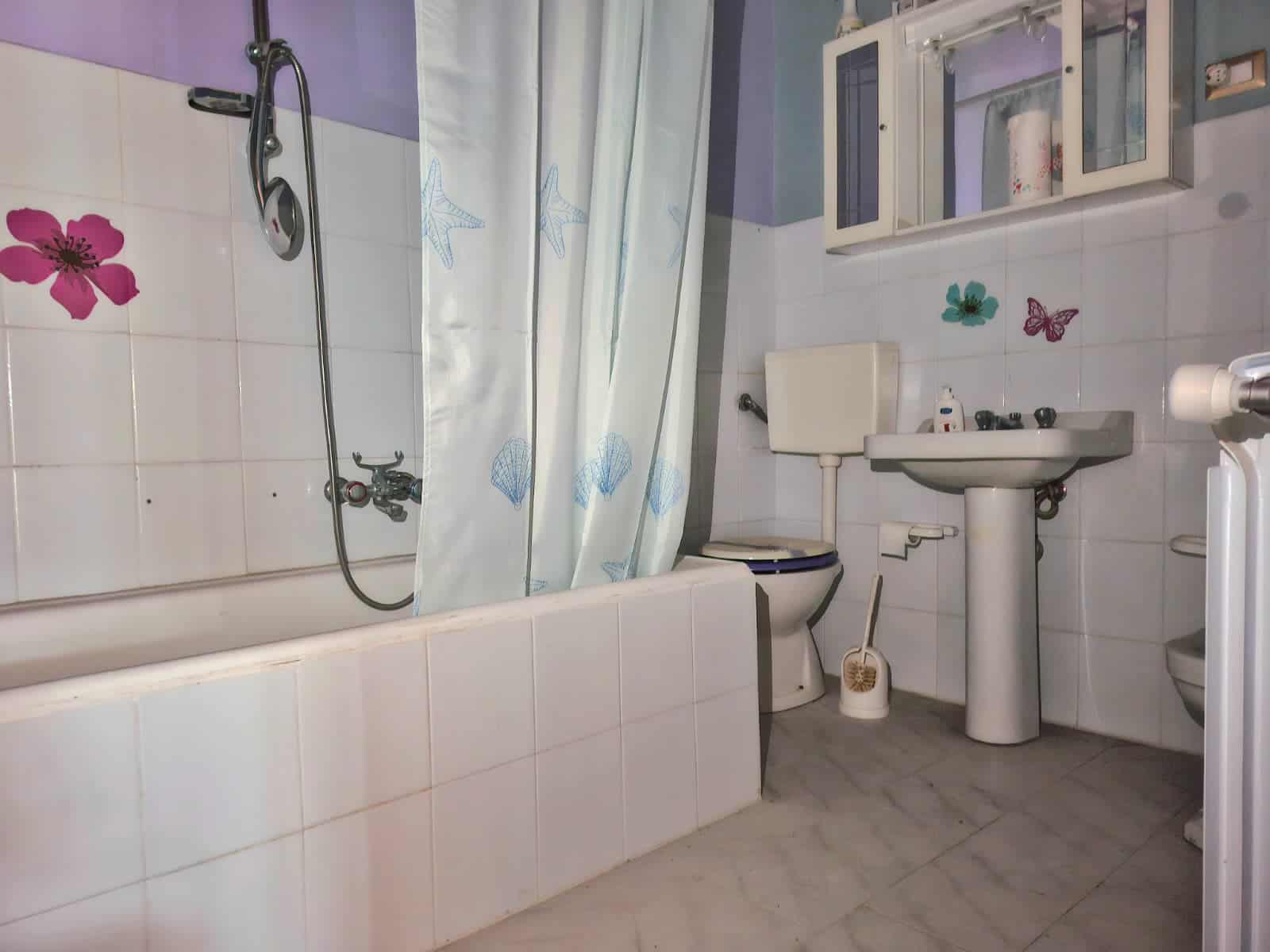 Stanza da bagno con piastrelle bianche, lavandino, wc e vasca con doccia e tenda. Decorazioni floreali e farfalle sulle pareti, armadietto sopra il lavabo. Ambiente funzionale e ben segnalato.