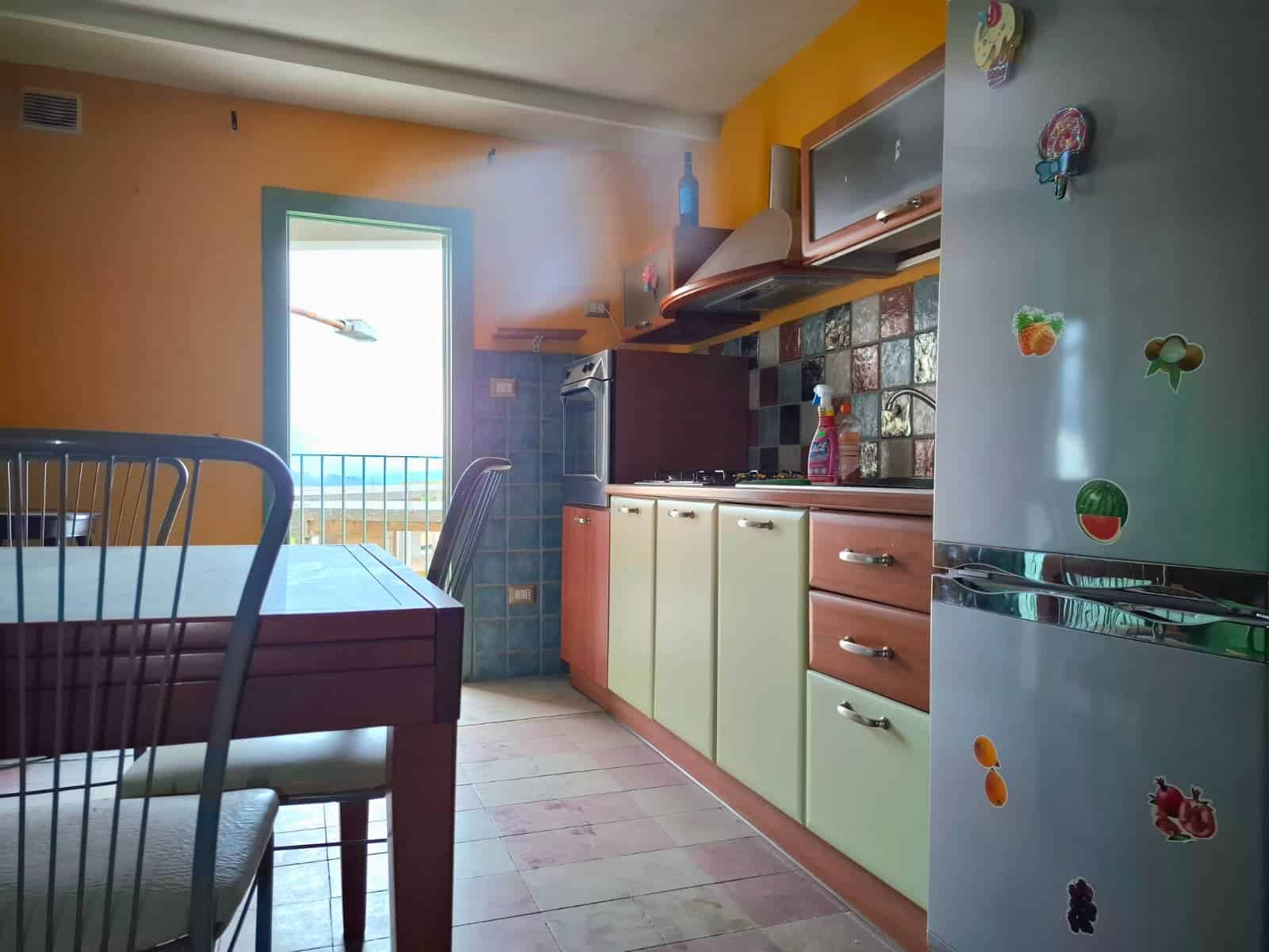 Estar cucina con mobili colorati, frigorifero decorato con stickers di frutta, tavolo e sedie, porta aperta su balcone con vista mare, ambiente luminoso e accogliente.