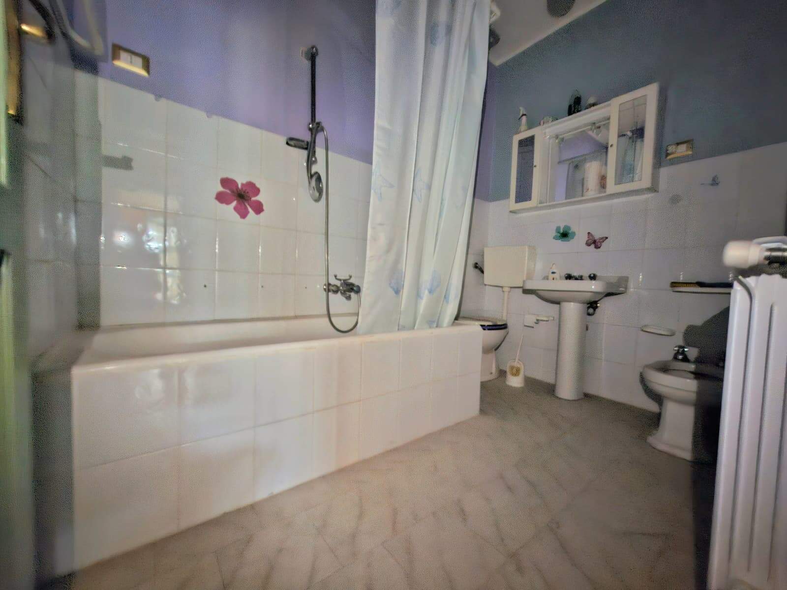 Bagno spazioso con vasca da bagno, lavabo, WC e bidet, arredato con piastrelle bianche e dettagli floreali, ideale per appartamenti in vendita o affitto, in zona centrale.