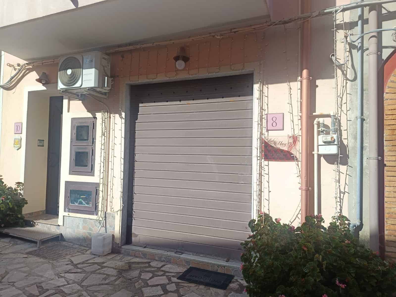 Persiana avvolgibile in alluminio grigio frontale con ingresso pedonale e sistema di illuminazione esterna, situata in una residenza moderna con decorazioni floreali e impianti di distribuzione esterni.