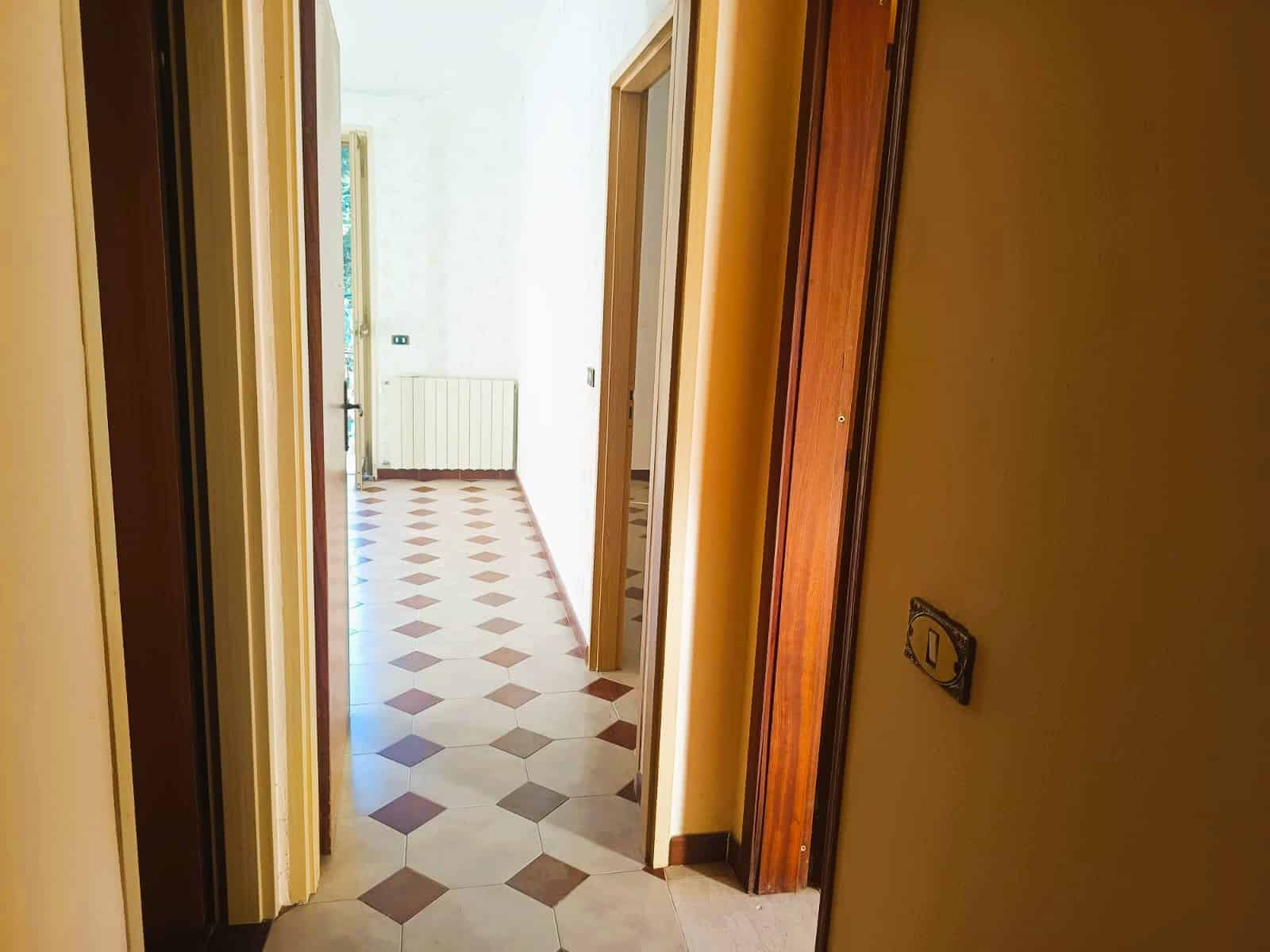 Vista di un corridoio luminoso e spazioso in un appartamento con pareti color crema e pavimento in piastrelle beige e marroni a scacchi, con porte in legno massiccio.