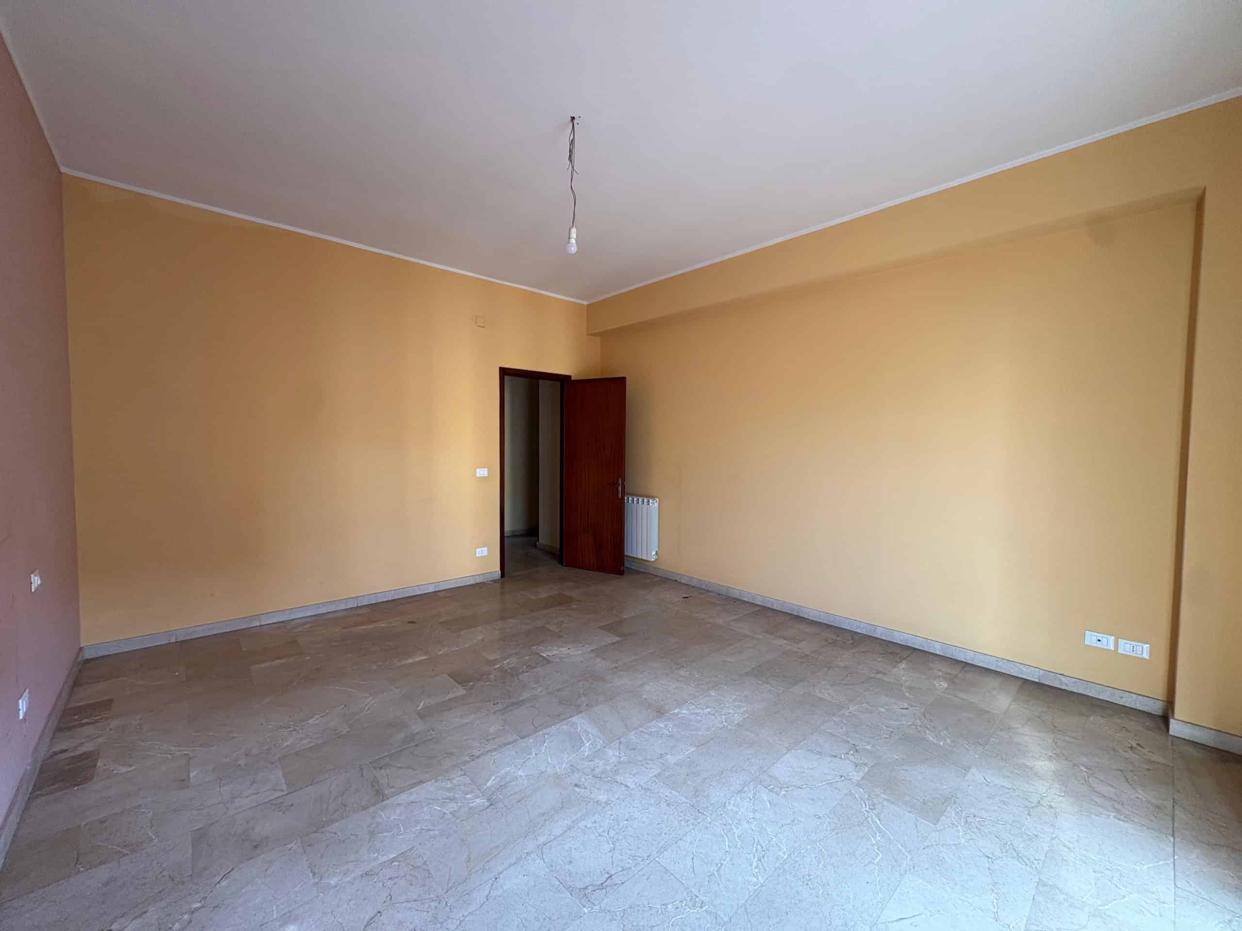 Ampio soggiorno vuoto con pareti color beige, pavimento in marmo e porta in legno, pronta per arredare. Ideale per trasformare in uno spazio confortevole e personalizzato.