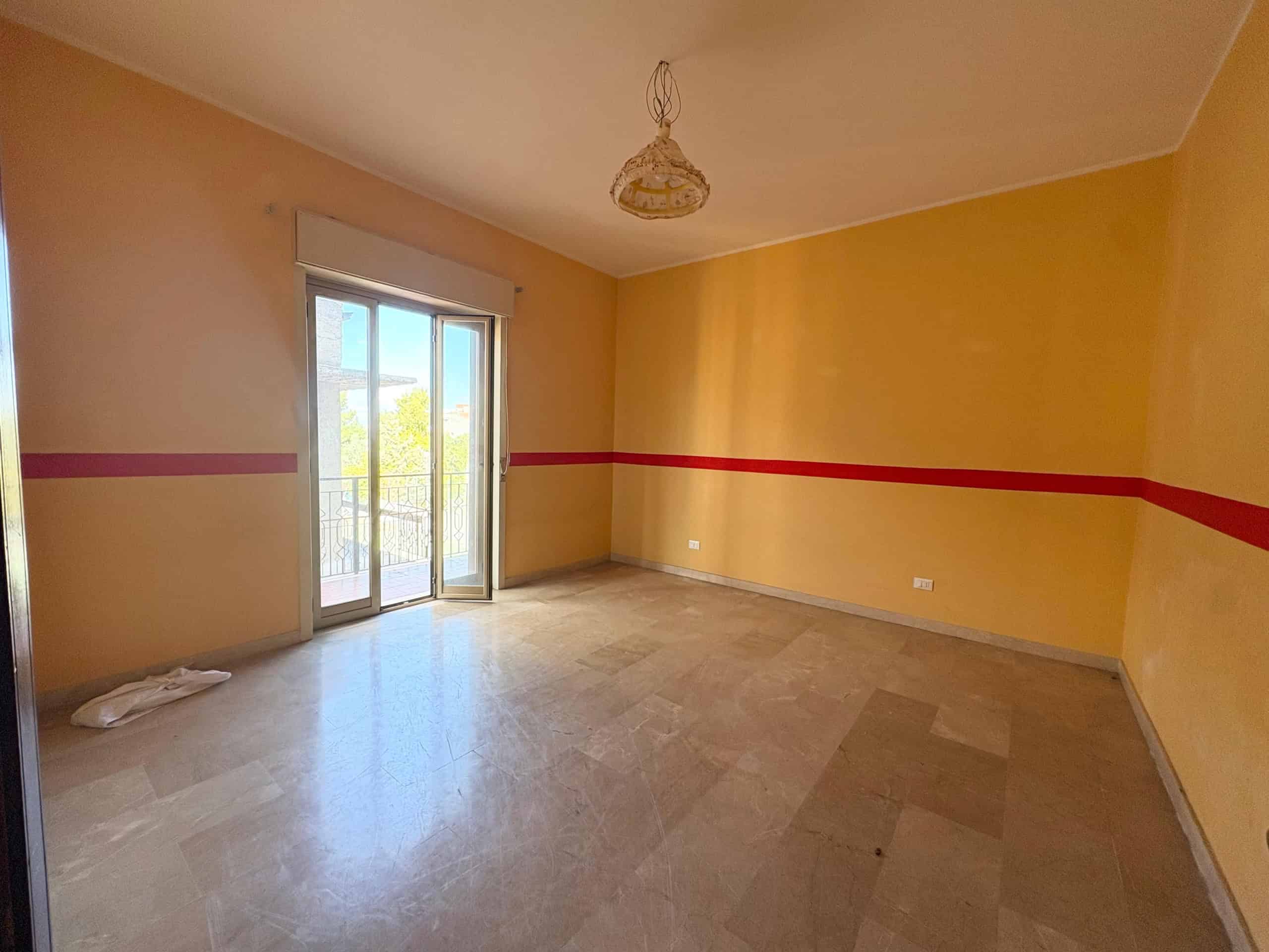 Ampio soggiorno con pareti gialle e dettaglio rosso, accesso a balcone con vista aperta, pavimento in marmo e luce naturale, ideale per abitazione o investimento immobiliare.