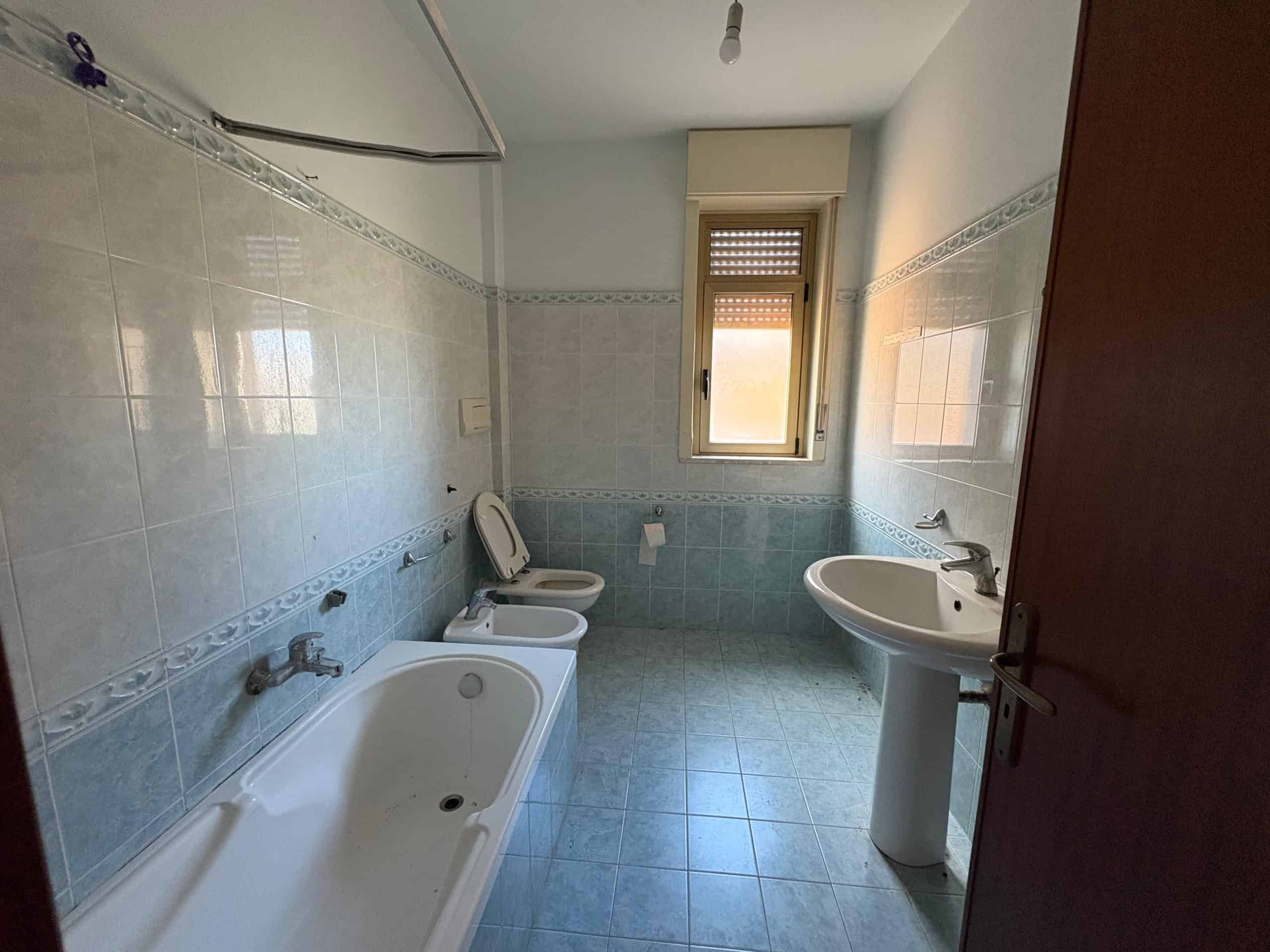 Bagno di casa in vendita con vasca, lavabo e WC in ambiente luminoso, finestra naturale, piastrelle azzurre e dettagli decorativi, ideale per ristrutturazione e affitto o residenza.