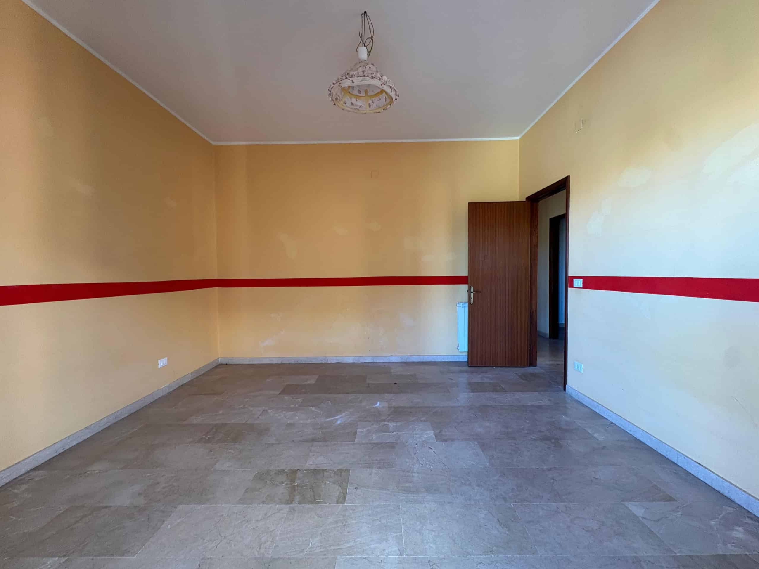 Spazio vuoto e luminoso in appartamento NextCasa, con pareti gialle e dettaglio rosso, pavimento in gres, porta in legno e luci in soffitto, ideale per ristrutturazione o arredo.