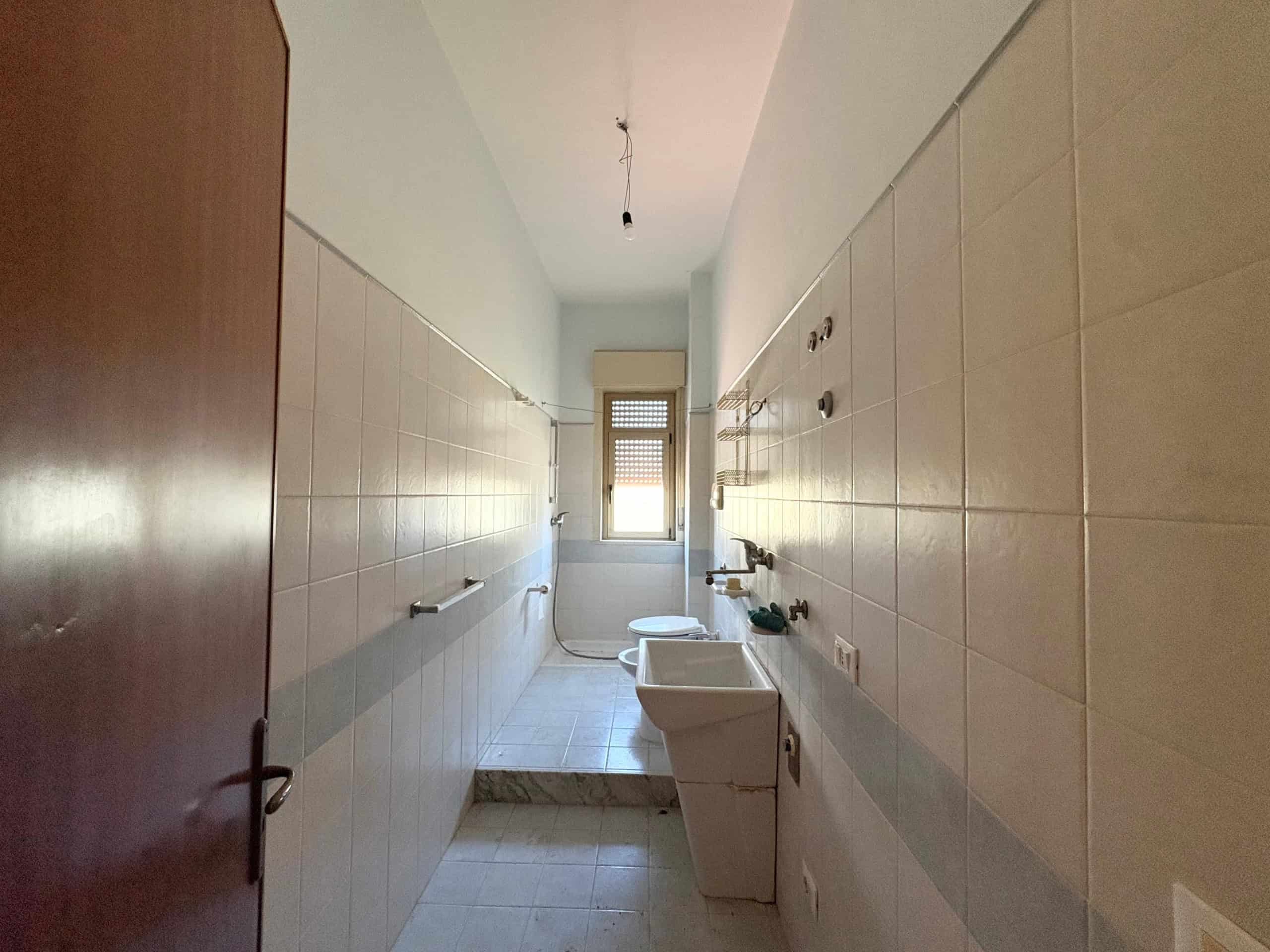 Ampio vano lavanderia con finestra, pareti rivestite di piastrelle beige, lavandino e spazio per lavatrice, luce naturale, ideale per rinnovamento e utilizzo funzionale.