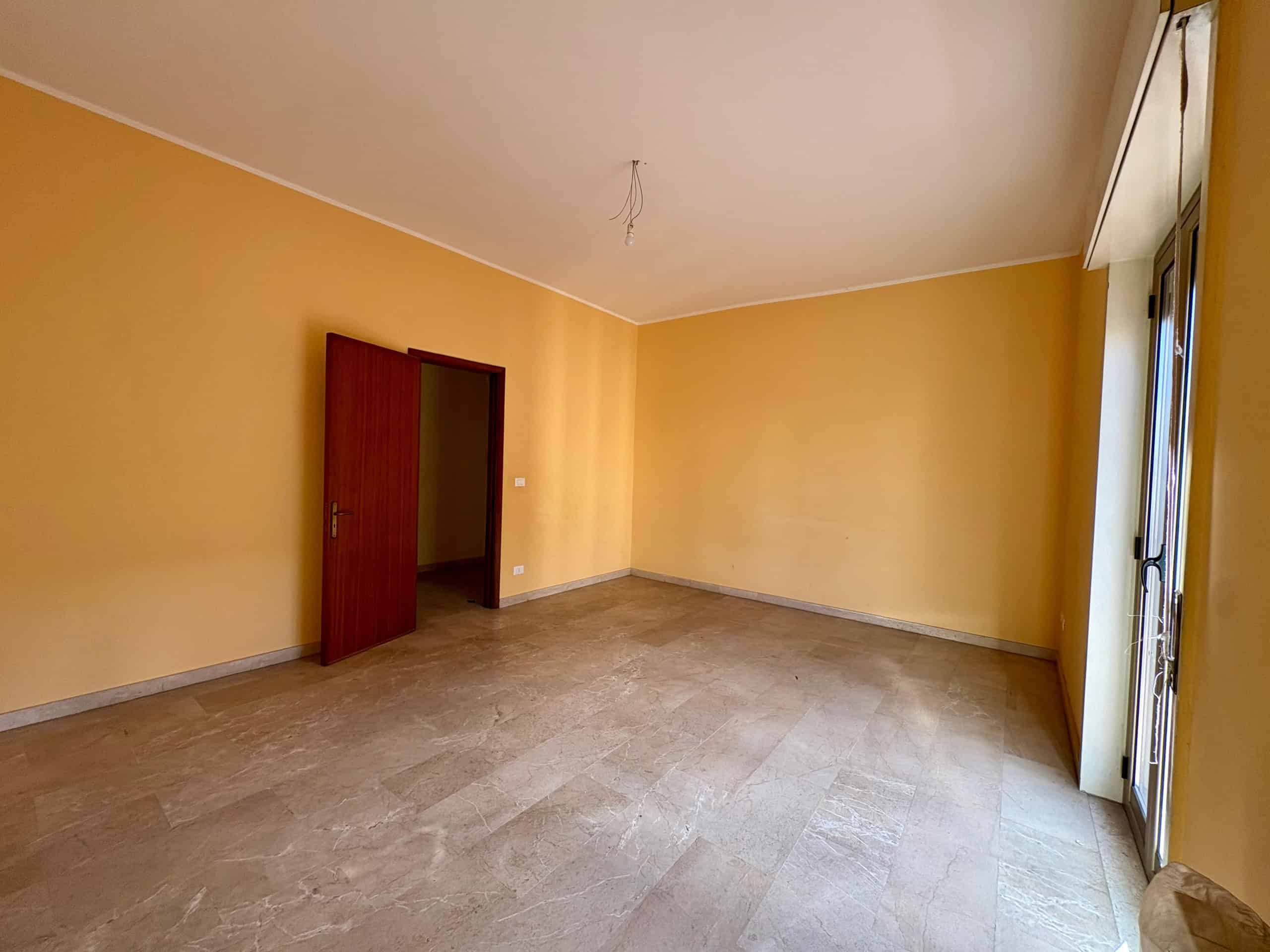 Ampio soggiorno con pareti gialle, pavimento in marmo, porta in legno e grande finestra con accesso al balcone, ideale per rilassarsi o ricevere ospiti in un ambiente luminoso e confortevole.