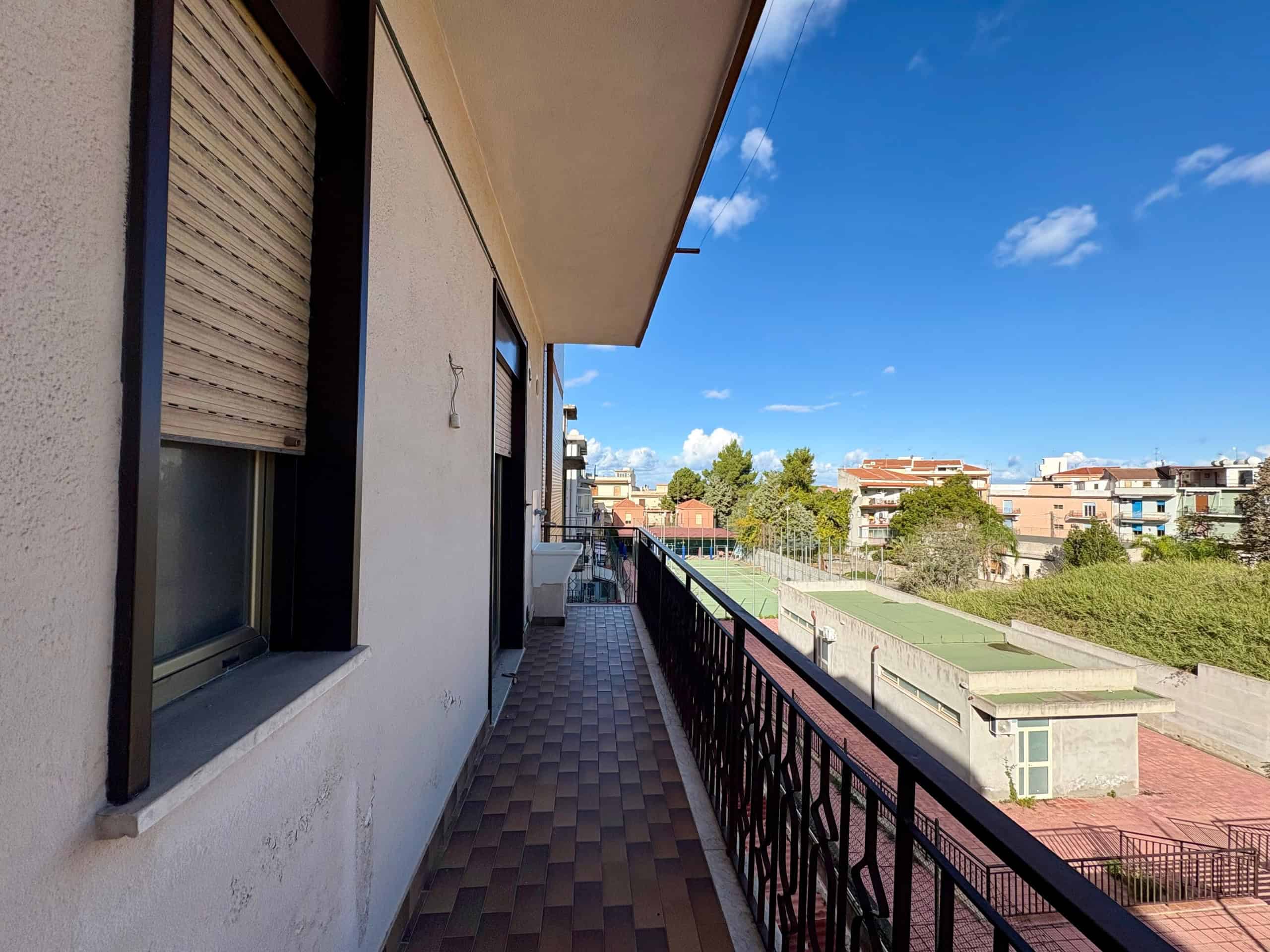 Balcone di un appartamento con vista sul campo da tennis, case residenziali e ampio skyline in lontananza, cielo azzurro e giornata soleggiata, ambiente silenzioso e tranquillo.