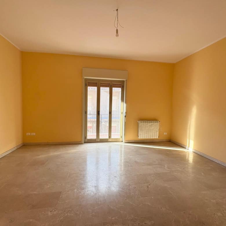 Ampia stanza vuota con pareti gialle e pavimento in marmo, luminosa con balcone e radiatore, ideale per sistemi di riscaldamento efficienti e luce naturale, perfetta per arredamenti moderni e accoglienti, NextCasa.