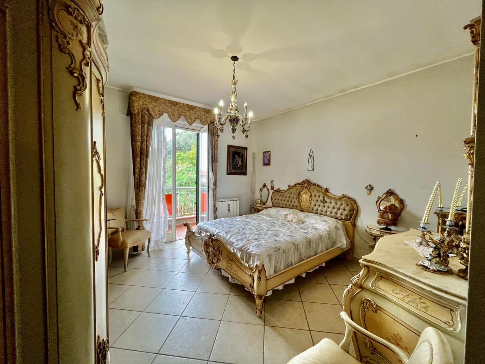 Elegante camera da letto in stile classico con mobili dorati e dettagli raffinati, con accesso a un balcone con vista verde, ambientazione ideale per case di lusso e immobili di prestigio.