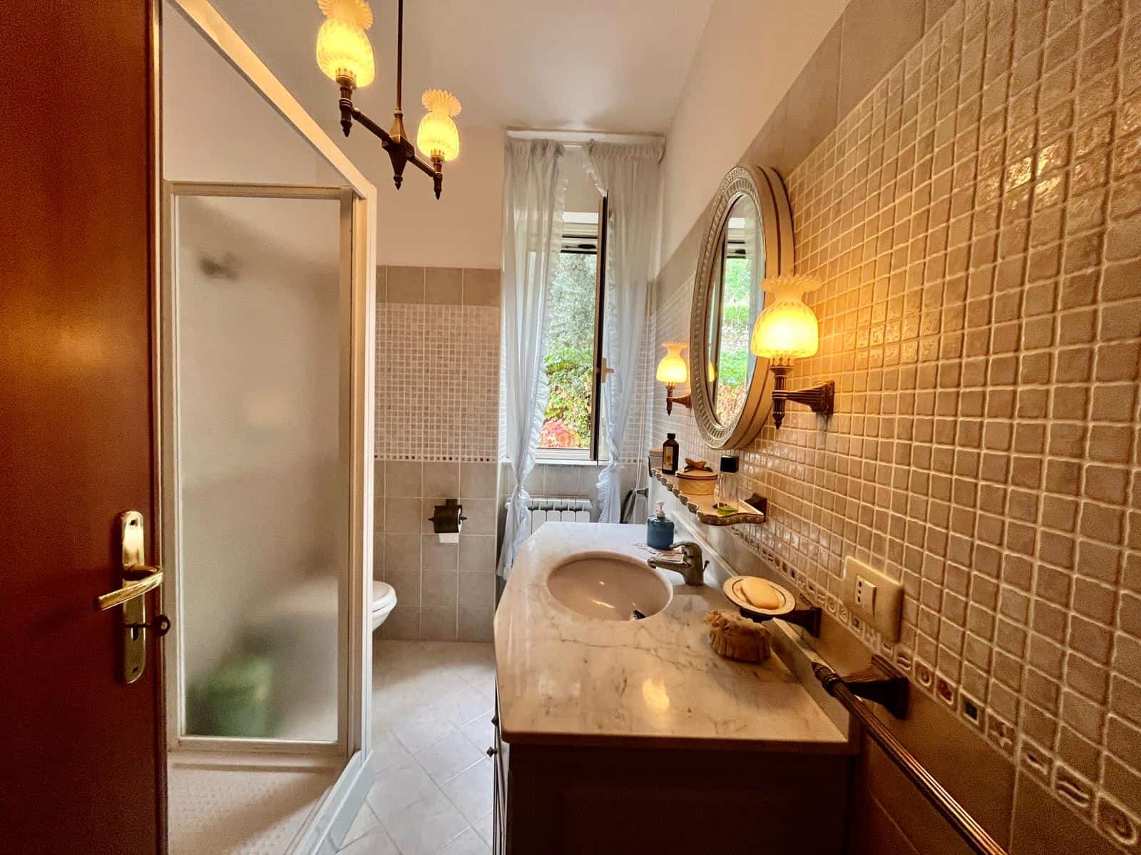 Bagno elegante e luminoso con piastrelle beige, lavabo in marmo, specchio rotondo, lampade vintage e finestra con tende leggere, ideale per la vendita o affitto di immobili di lusso.