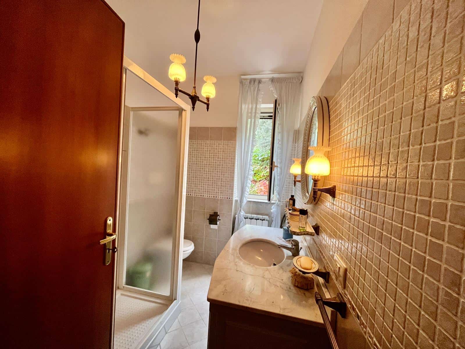Bagno classico con lavabo in marmo, pareti rivestite in mosaico beige e specchio rotondo, illuminato da applique vintage e luce naturale dalla finestra.